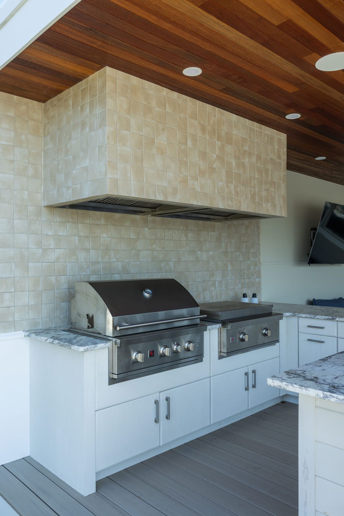 Modern outdoor kitchen with built-in grill, white cabinets, stone backsplash, and wood ceiling