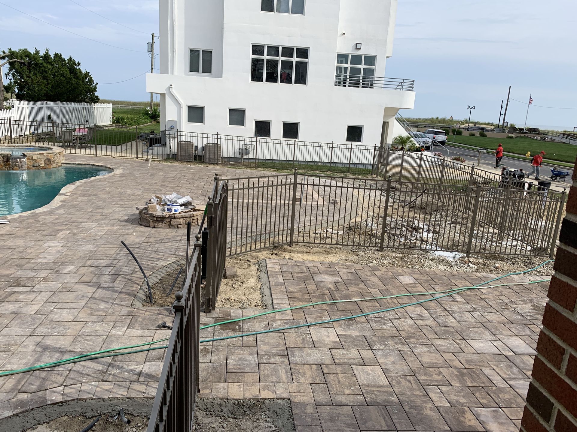 Patio with brick pavers and black railing beside a white seaside building and pool