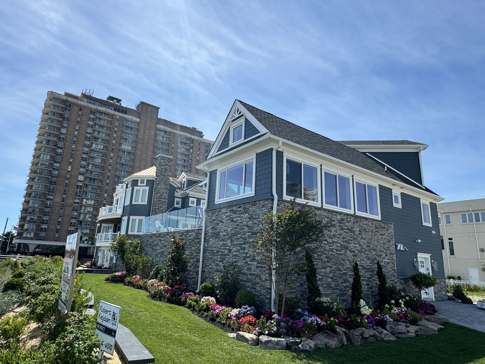 Stone house with white trim and flower garden beside a tall apartment building under a blue sky