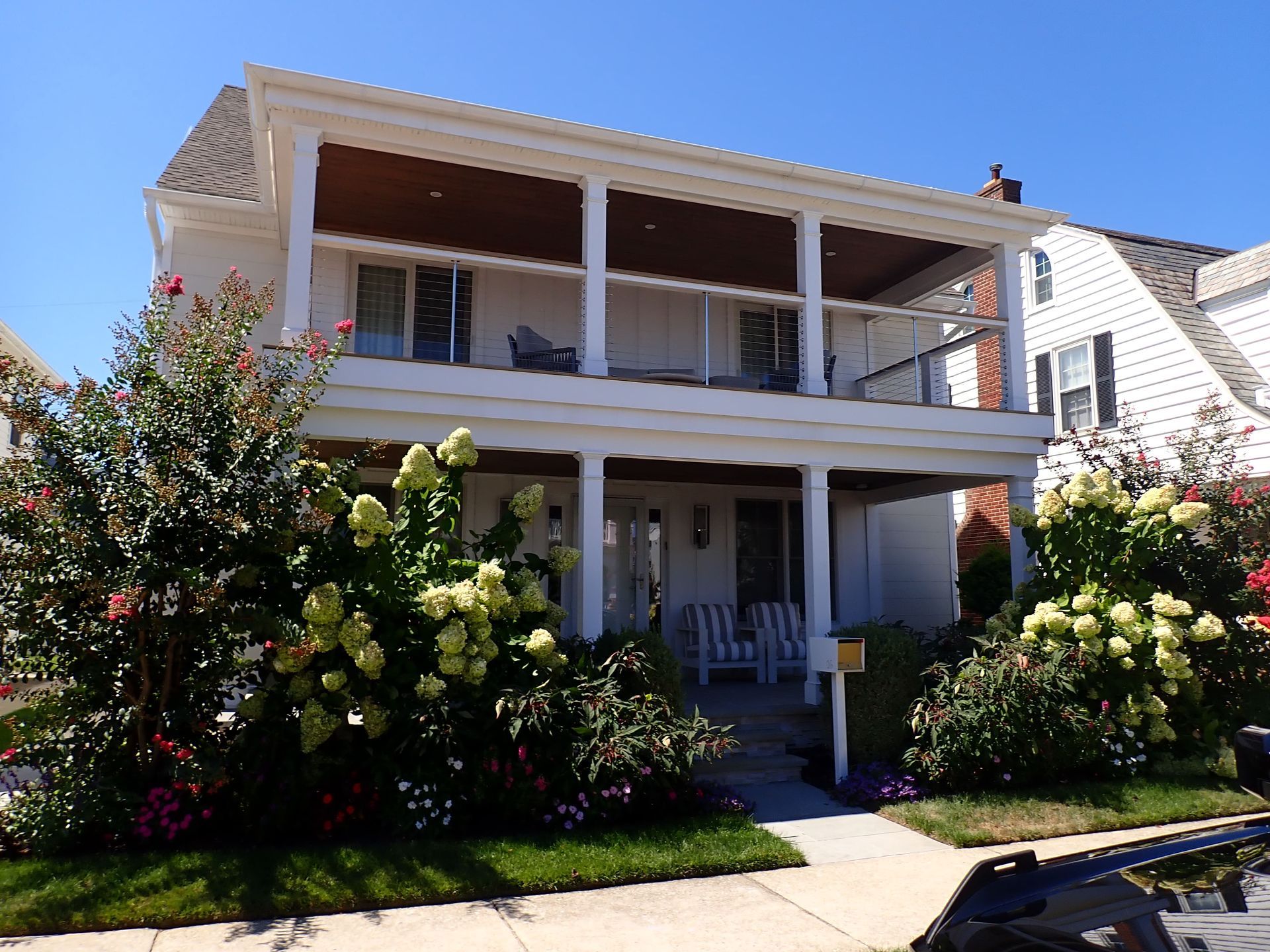 Two-story white house with wraparound balcony, lush front garden, and a shaded porch