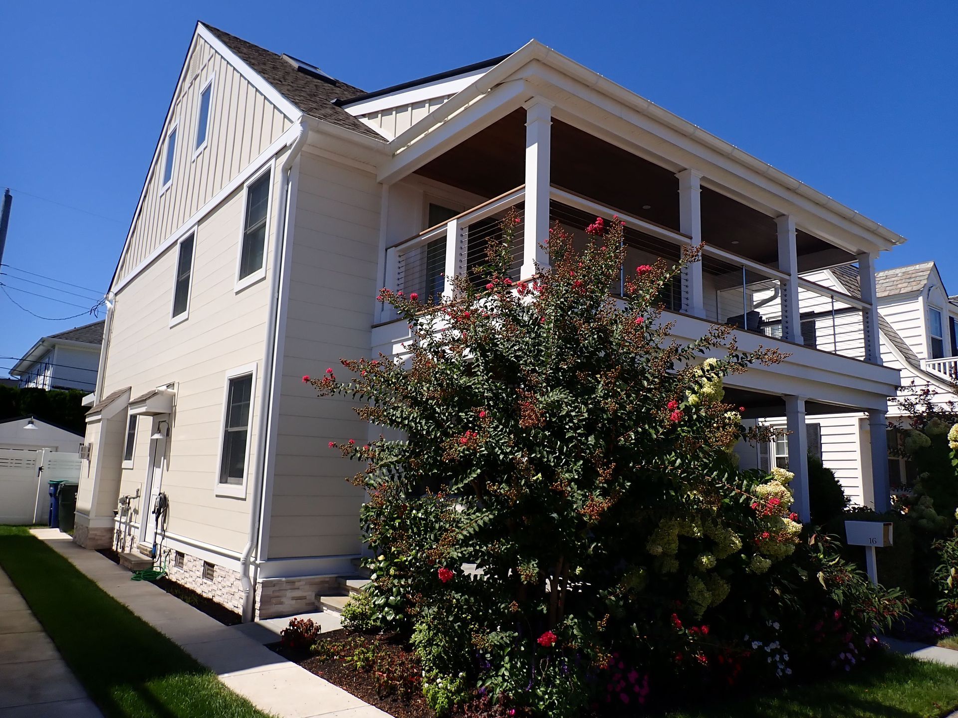 Two-story beige house with a covered porch and flowering bush in front under a clear blue sky