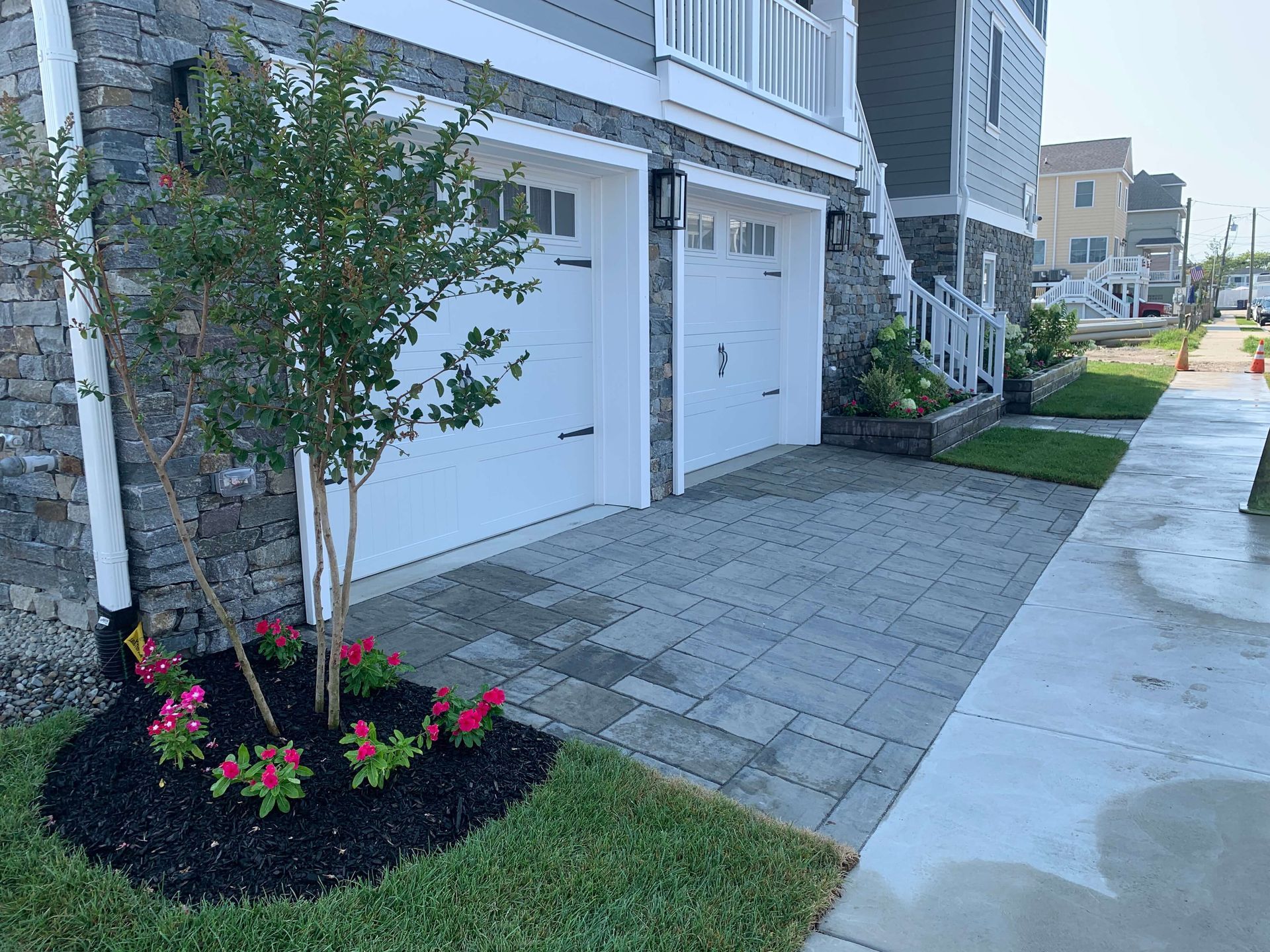 Townhouse driveway with white garage doors, paved walkway, and a small landscaped flower bed.