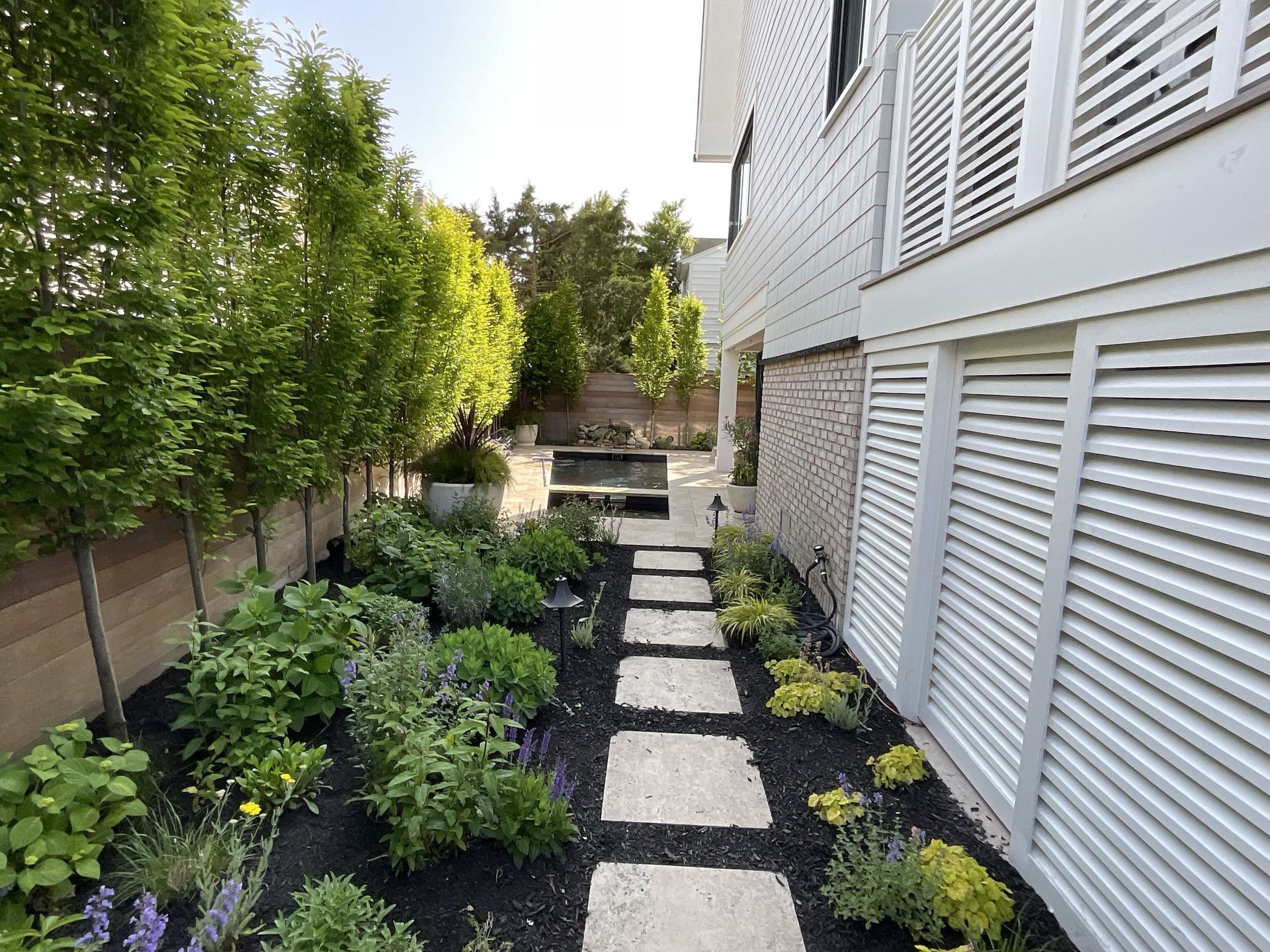 Narrow landscaped side yard with stepping stones, shrubs, and a house with white shutters on the right.