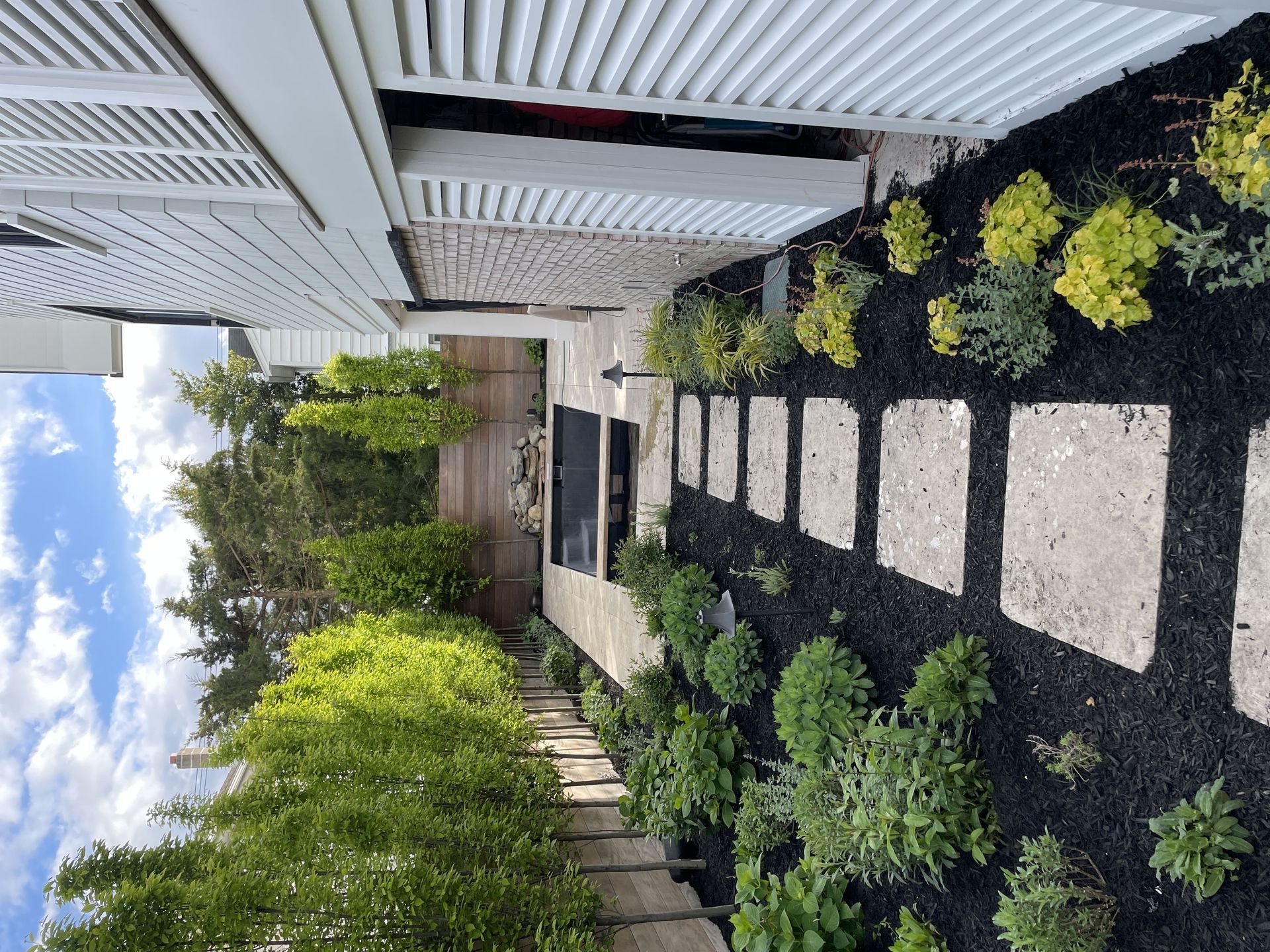 Narrow backyard walkway with stepping stones, greenery, and a white fence beside a house under a corrugated roof