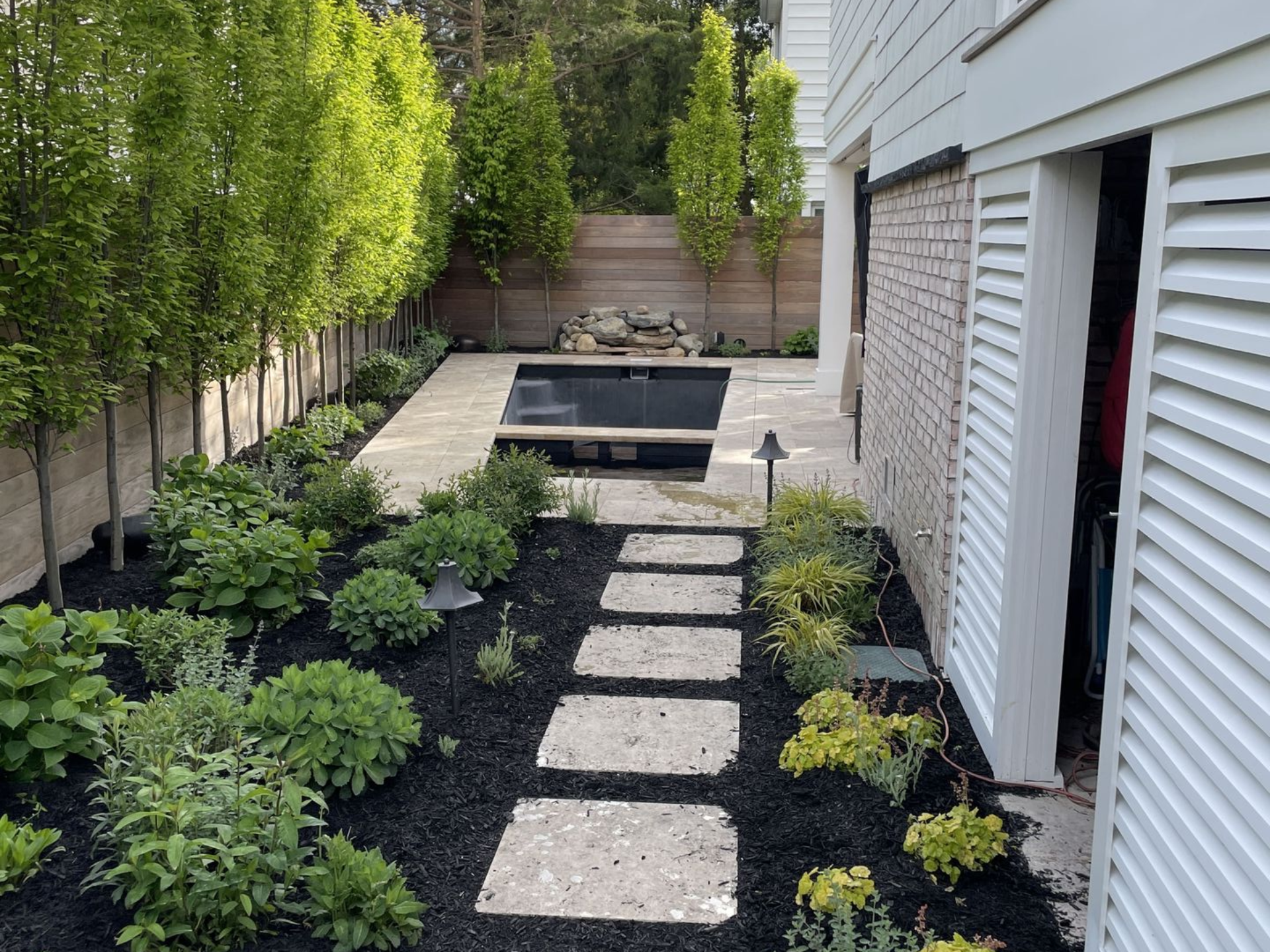 Narrow backyard path with stepping stones, black mulch beds, and a small rectangular pond beside a white house
