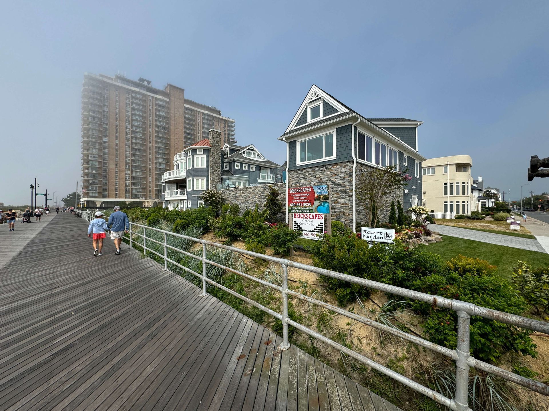 Boardwalk beside seaside homes and a tall apartment building under a hazy sky, with two people walking.