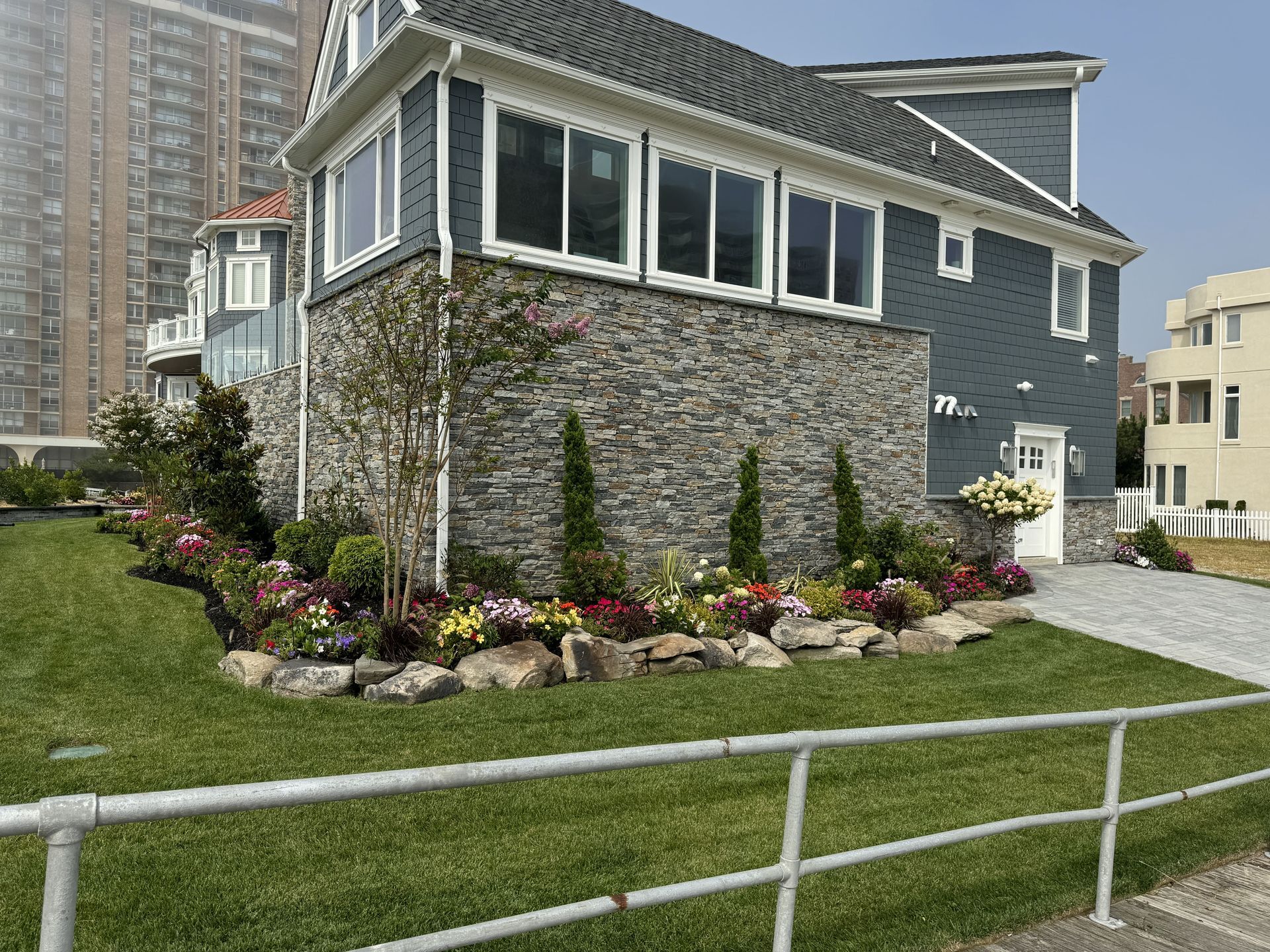Gray house with stone lower walls and flower garden beside a grassy lawn and white railing