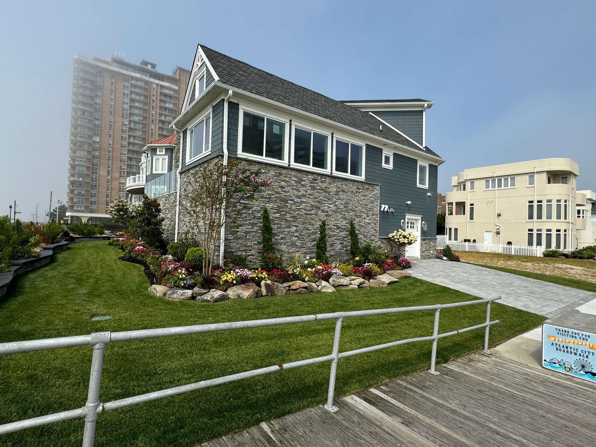 Gravel-roofed stone cottage on a grassy hill with flower beds, near a boardwalk and modern buildings.