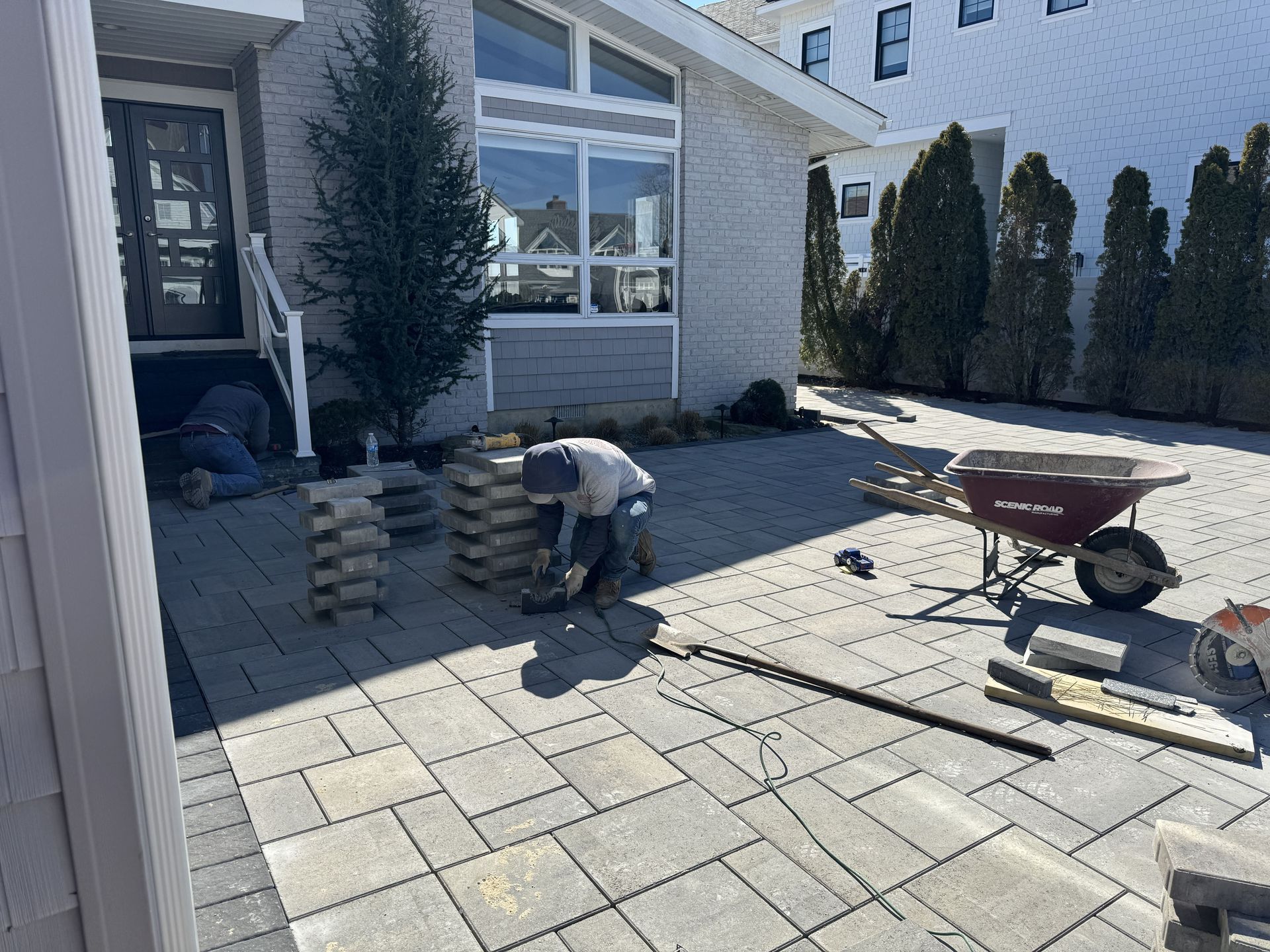 Person laying patio pavers in a front yard beside a house, with a wheelbarrow and stacked stones nearby.