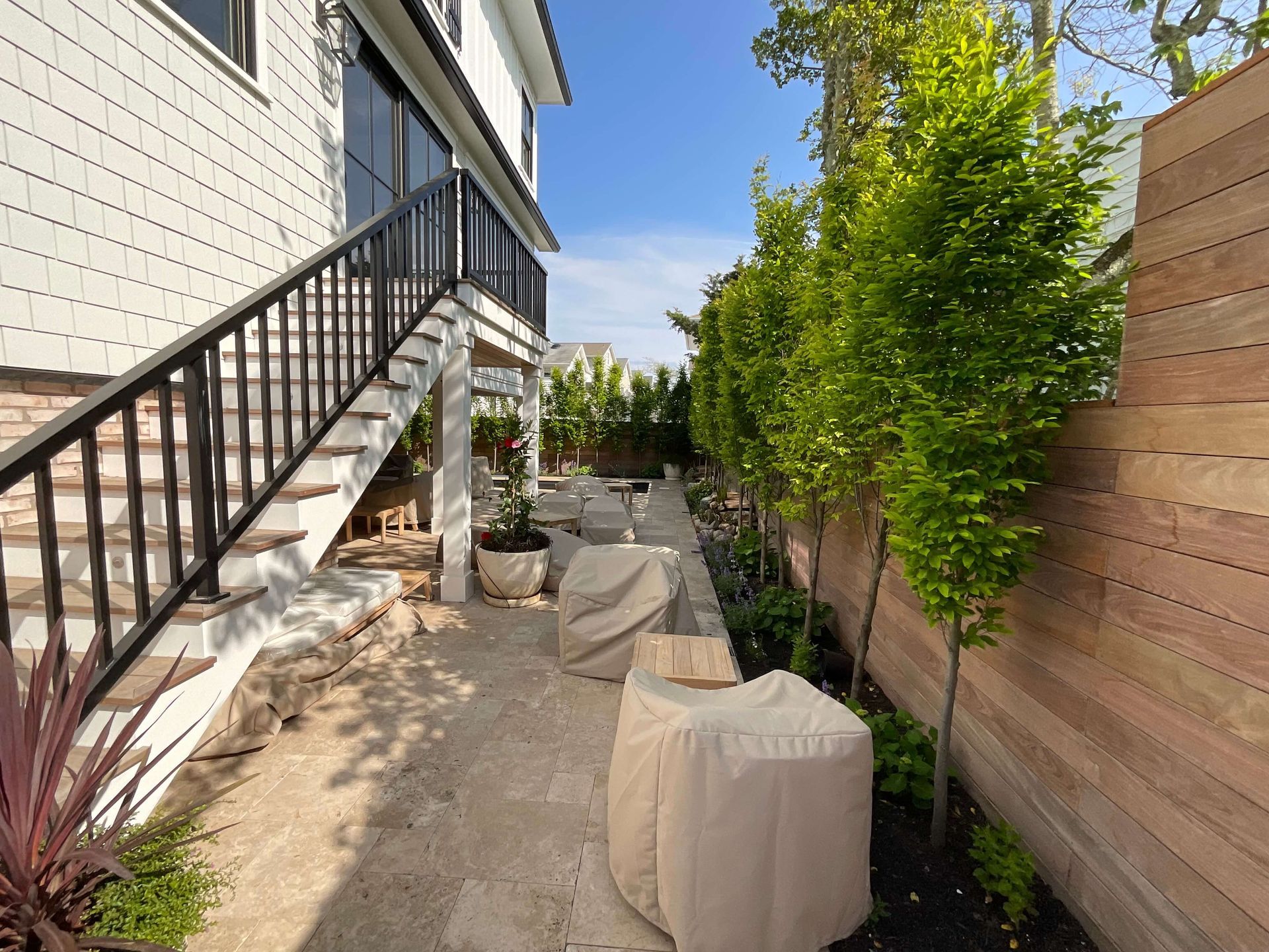 Narrow side yard with stairs, covered patio furniture, potted plants, and a wooden privacy fence along the house.