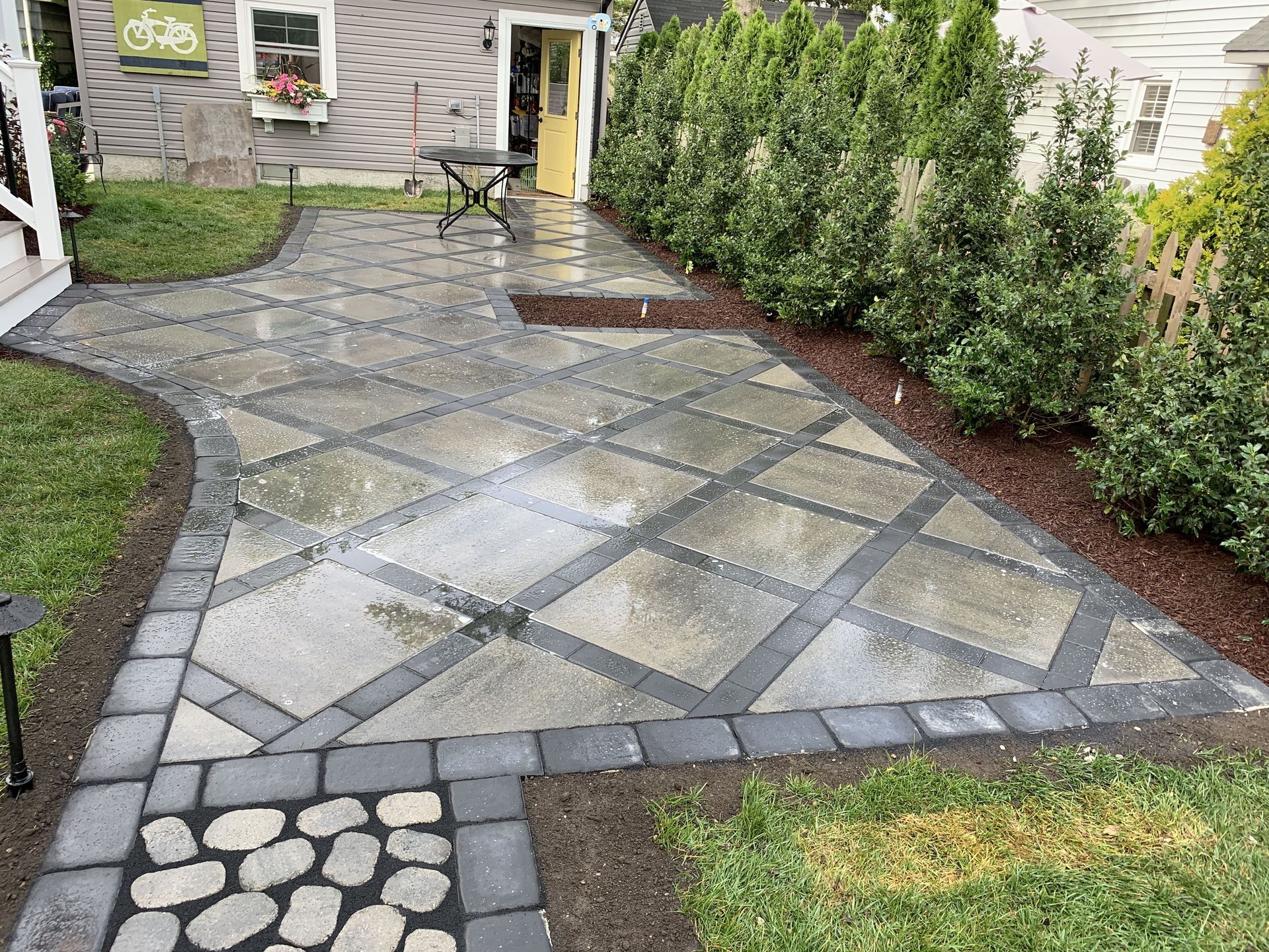 Paver patio with diagonal stone pattern beside shrubs and a small lawn in front of a house