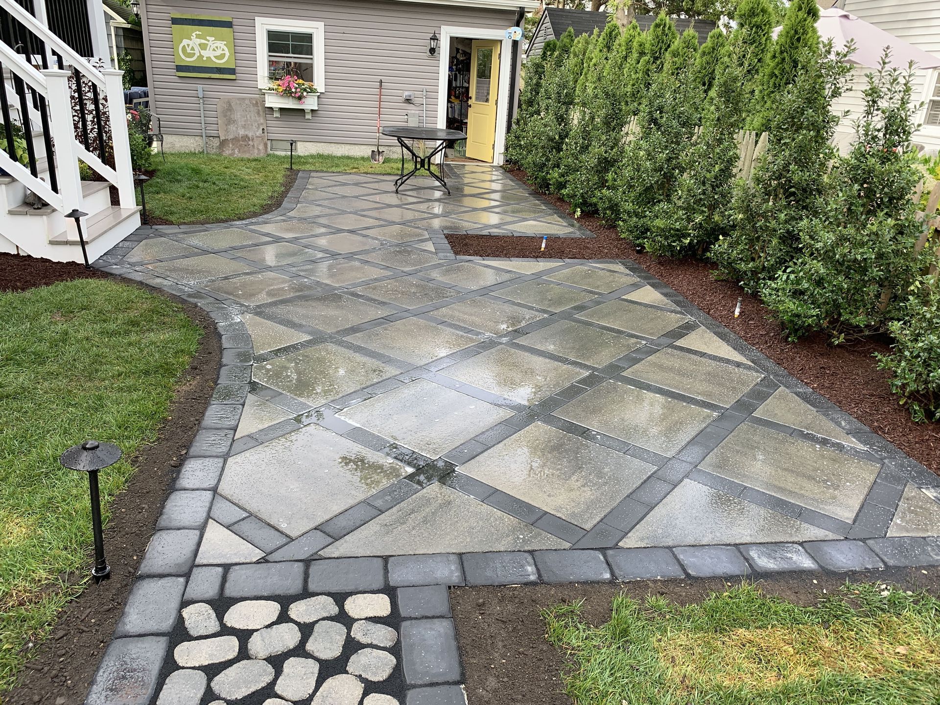 Stone-paver walkway leading to a house, bordered by shrubs and a small stepping-stone path.