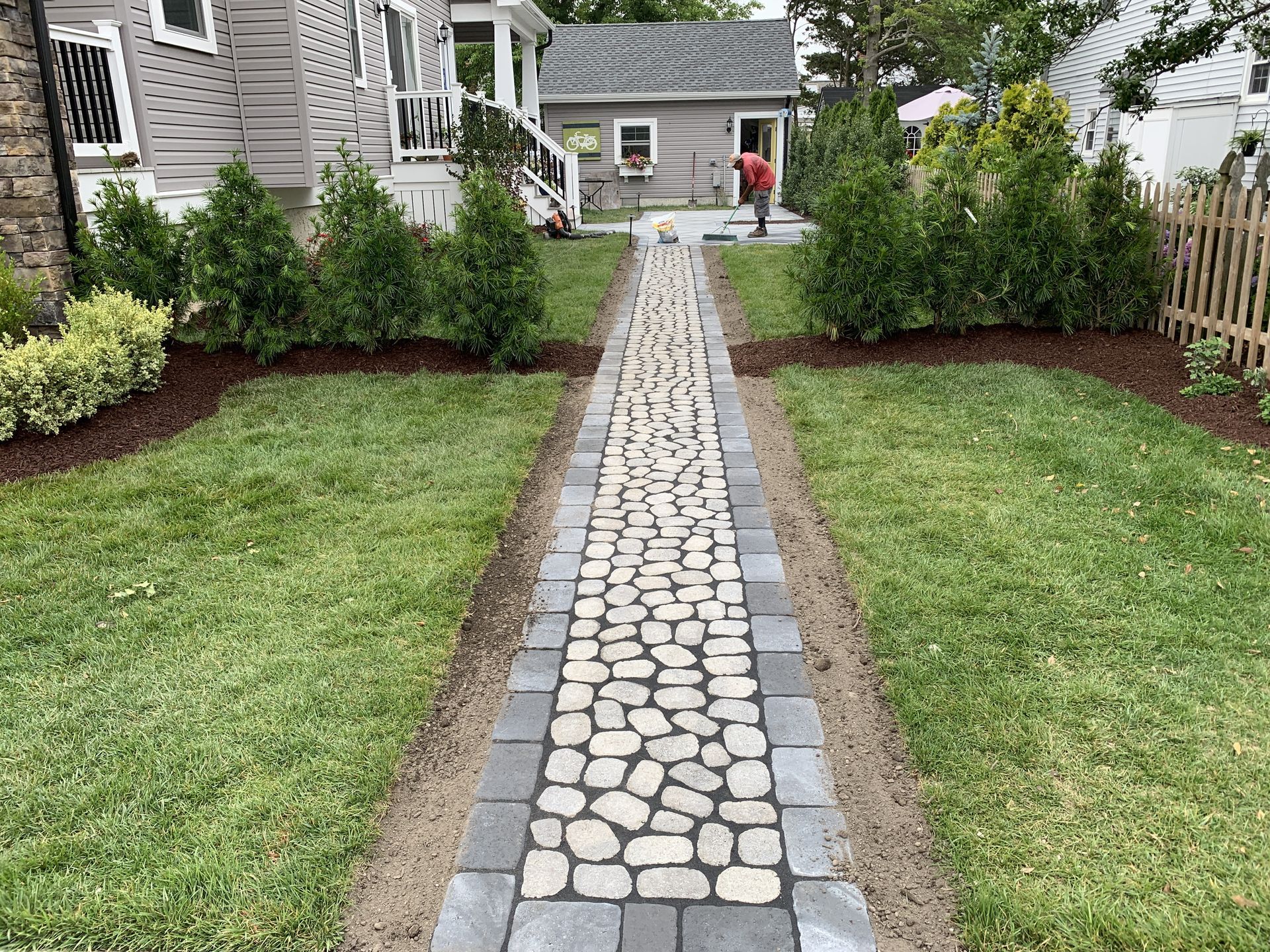 Stone walkway leading to a house, bordered by trimmed shrubs and lawn, with a person standing near the porch.
