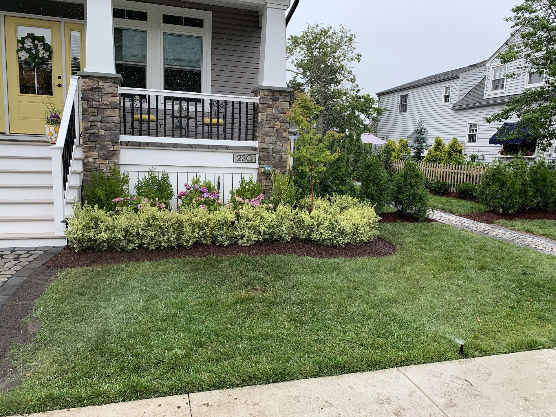 Front yard with green lawn, stone porch, and flowering shrubs beside a white house