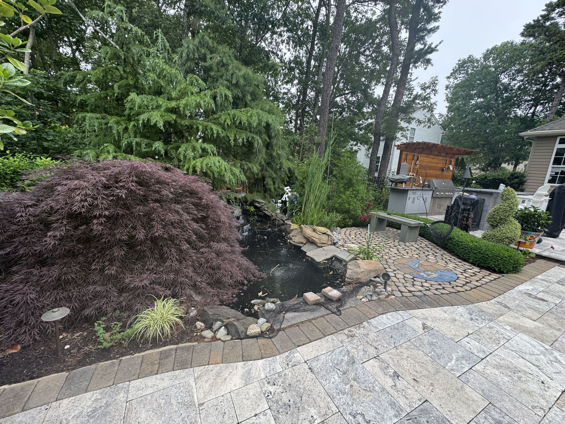 Landscaped garden with stone patio, shrubs, trees, and a small pond with waterfall