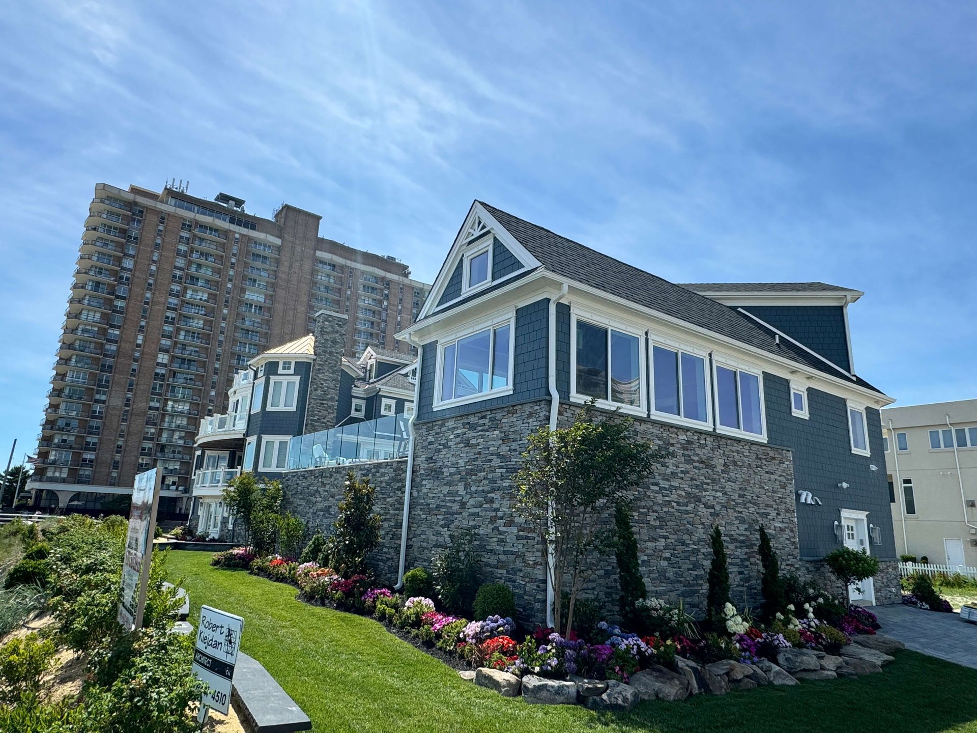 Stone house with blue-trimmed windows and flower beds beside a tall apartment building under a blue sky