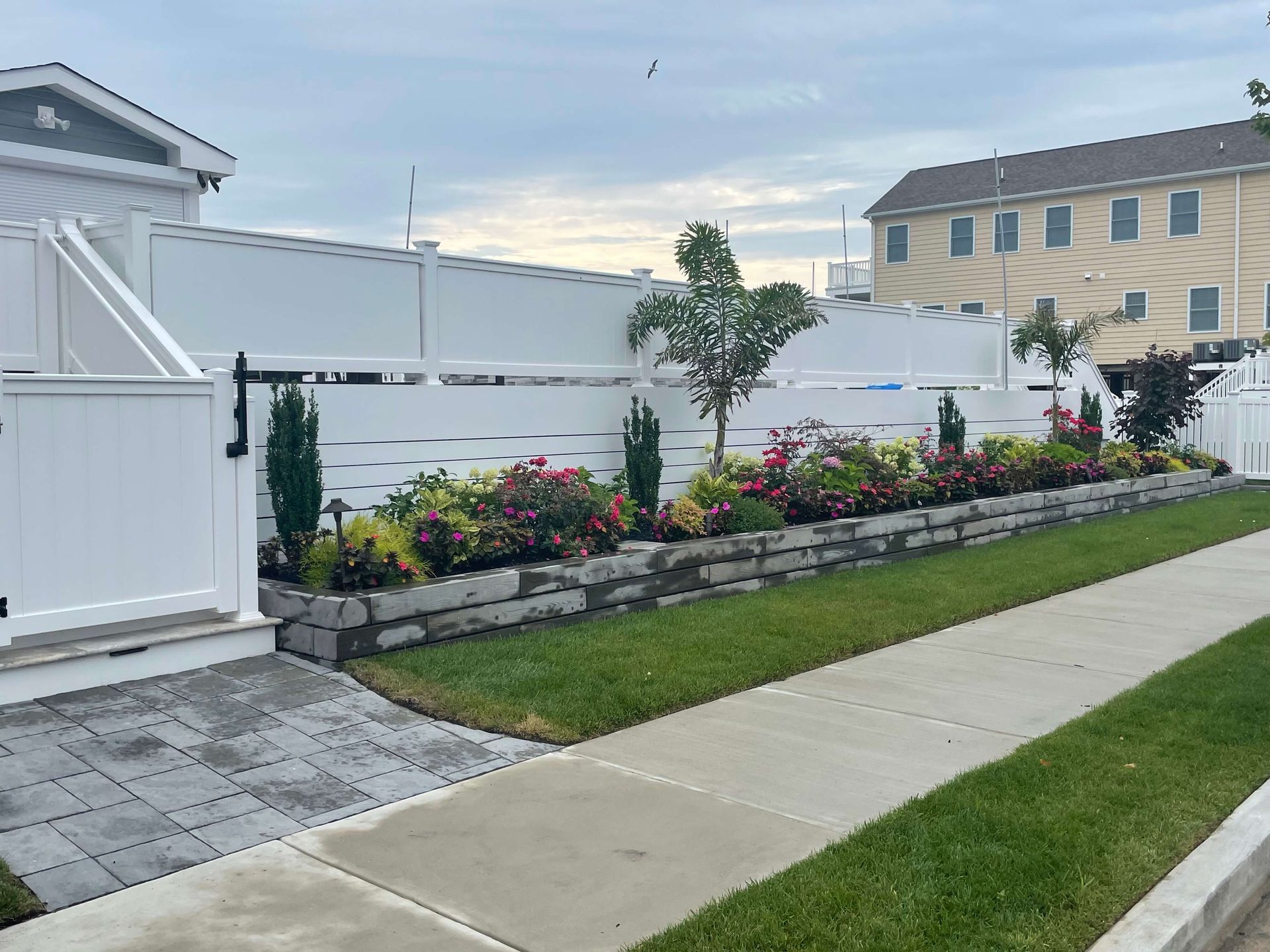Landscaped front yard with white fence, flower beds, and a sidewalk beside green lawn
