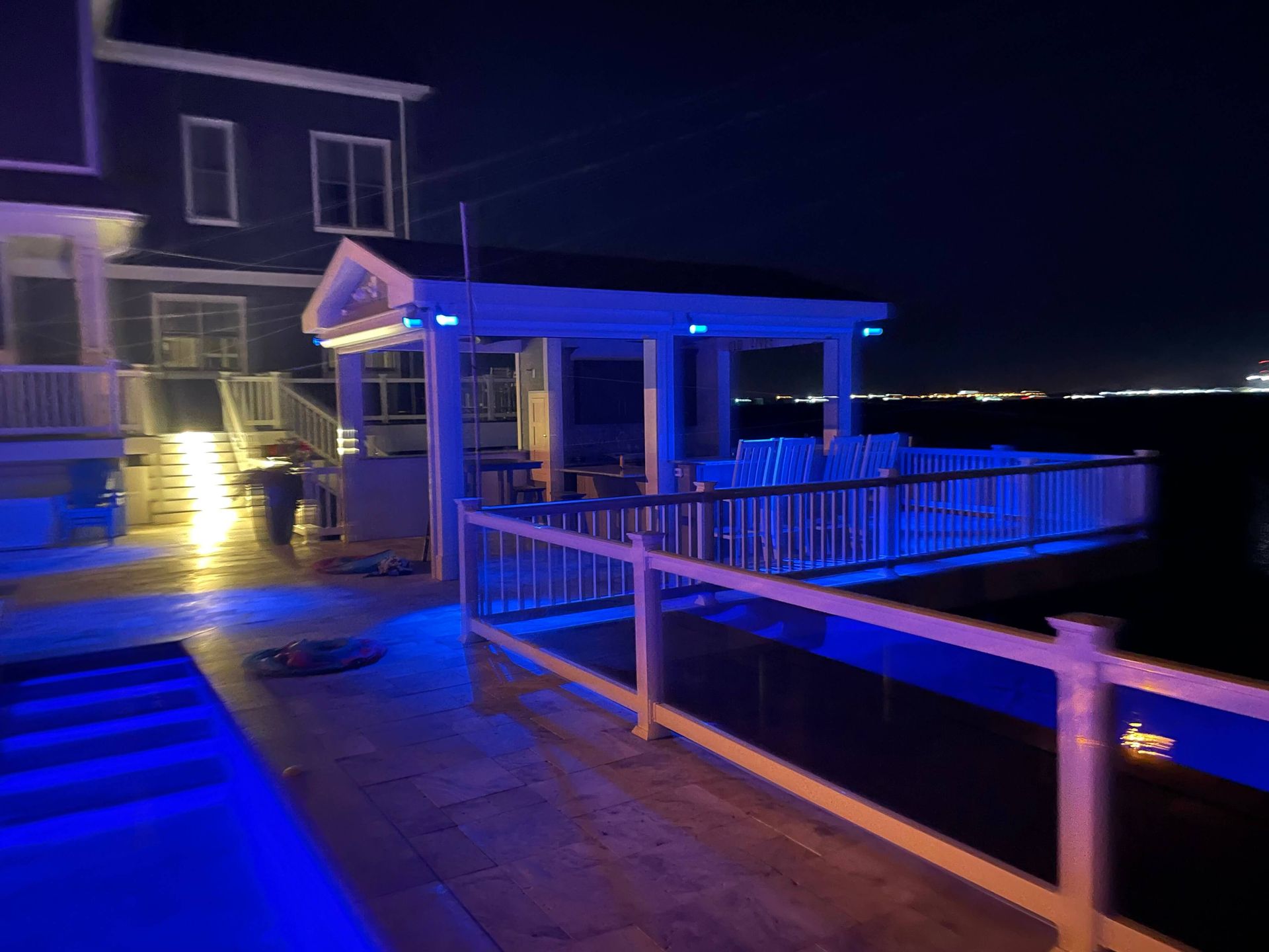 Blue-lit waterfront deck at night with railing, stairs, and lit house beside dark water