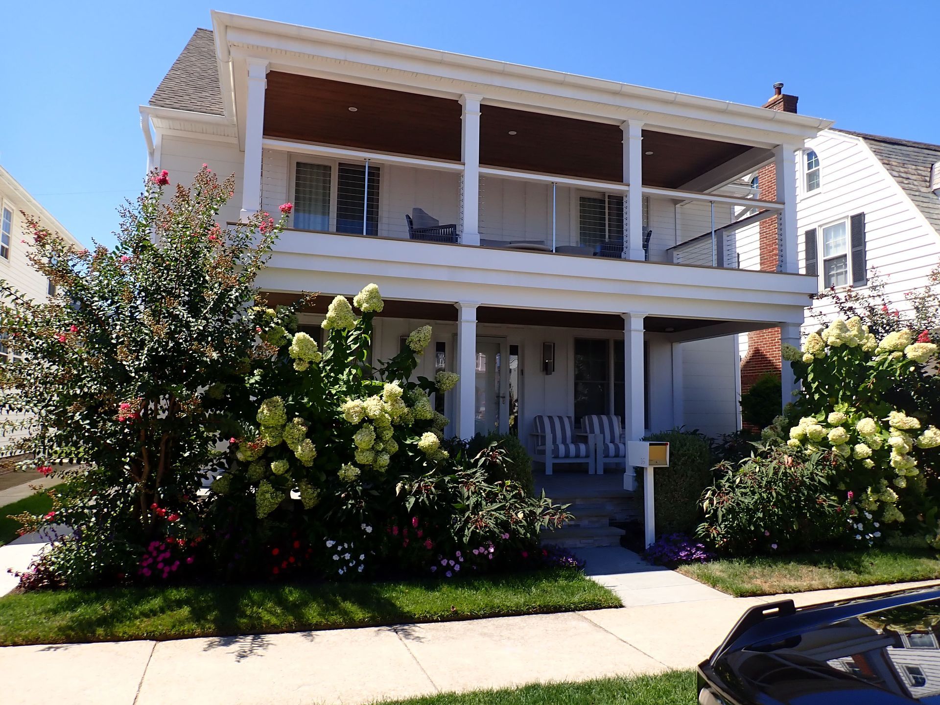 Two-story white house with a front porch, surrounded by lush flowering shrubs on a sunny day