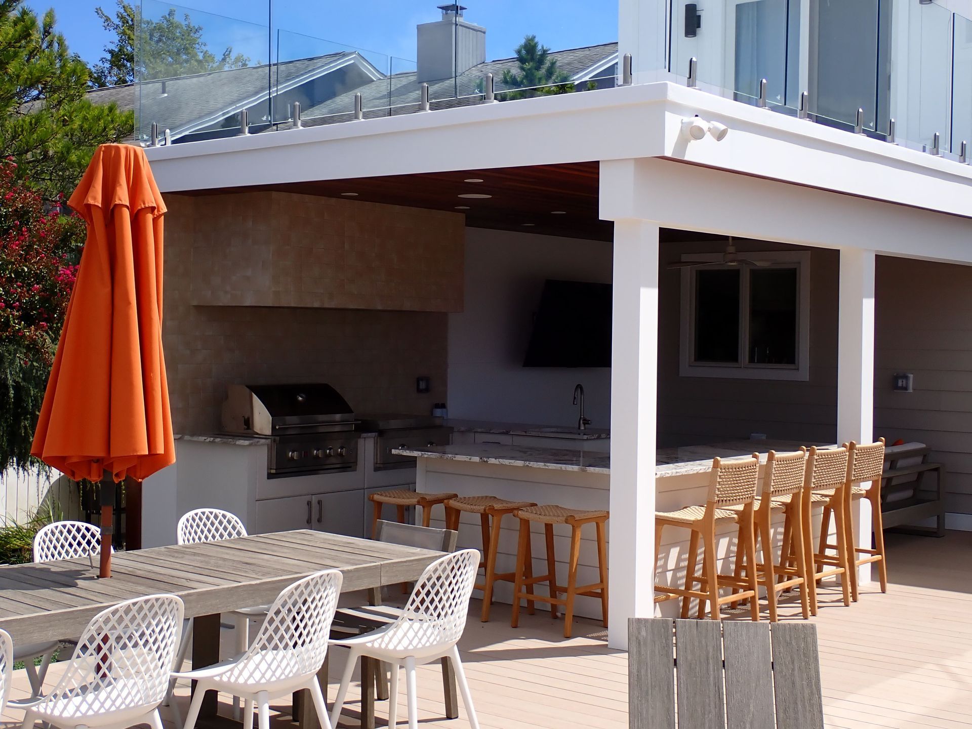 Outdoor patio with white tables and chairs, orange umbrella, bar stools, and a built-in grill under a white pergola.