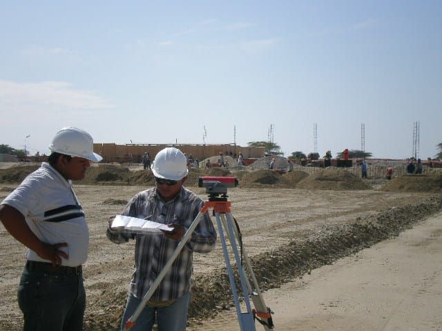 Dos trabajadores con cascos de seguridad utilizando un trípode de topografía en una obra de construcción.