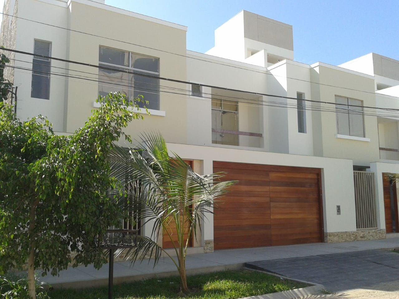 Moderna casa blanca de dos plantas con puerta de garaje de madera, árboles y entrada pavimentada.
