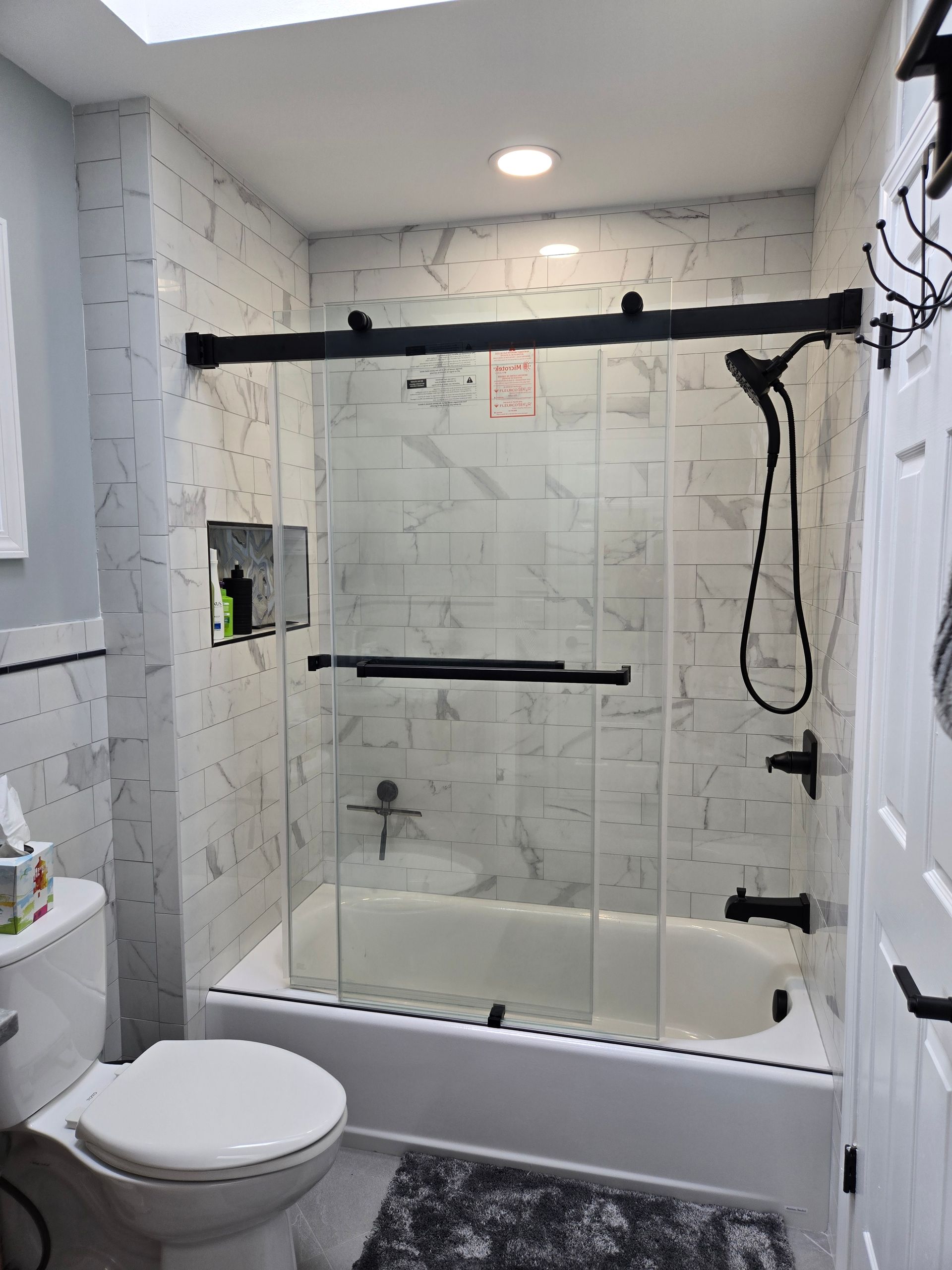 Bathroom with glass-enclosed shower over tub, black fixtures, white tiled walls, and toilet in foreground
