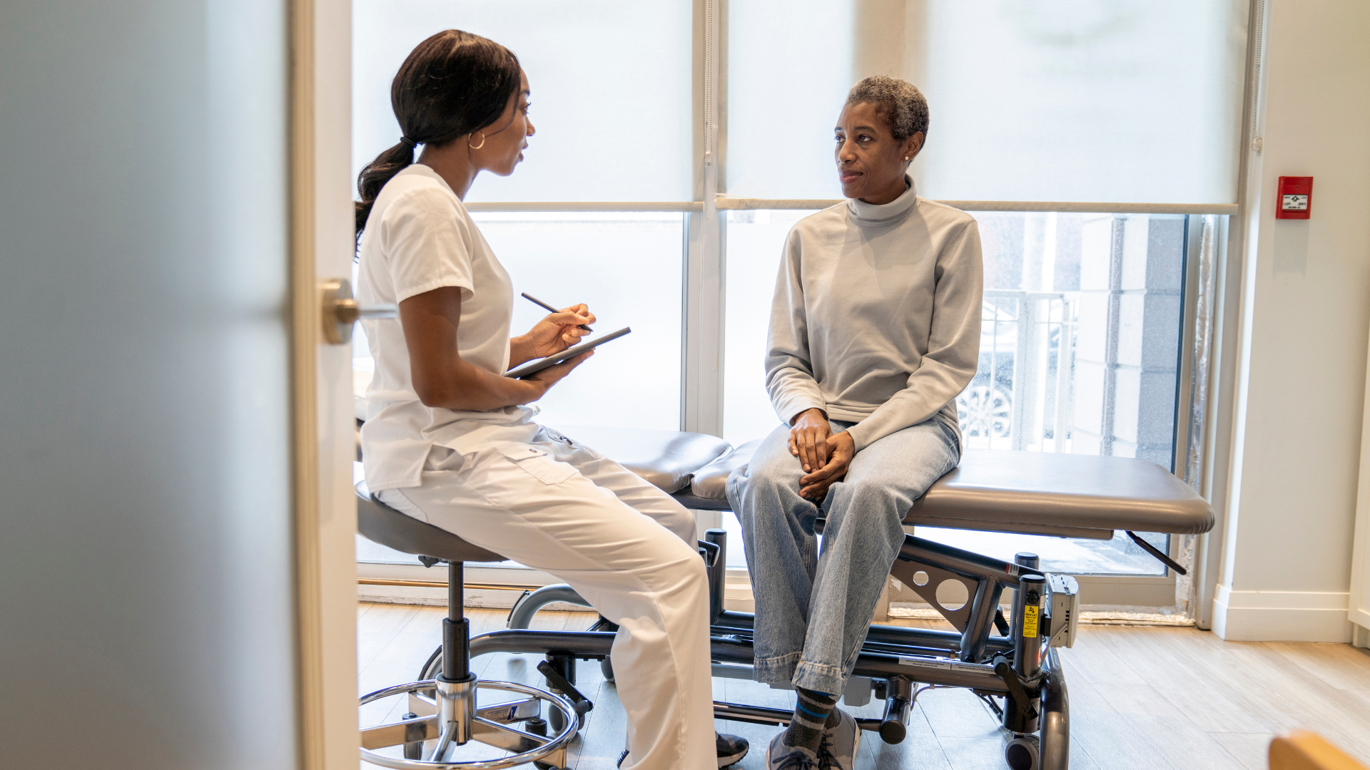 A healthcare professional consults with a patient in an exam room; the healthcare worker is taking notes on a tablet.