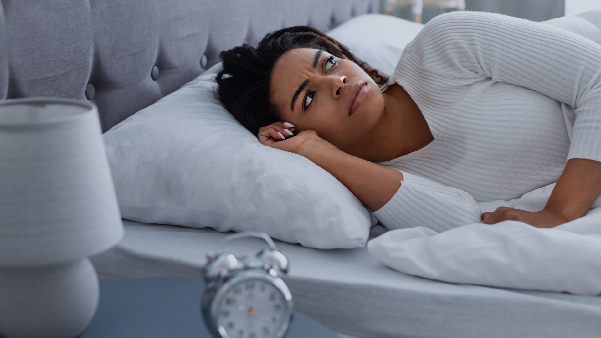 A person lies awake in bed at night, looking toward an alarm clock on the bedside table with a concerned expression.