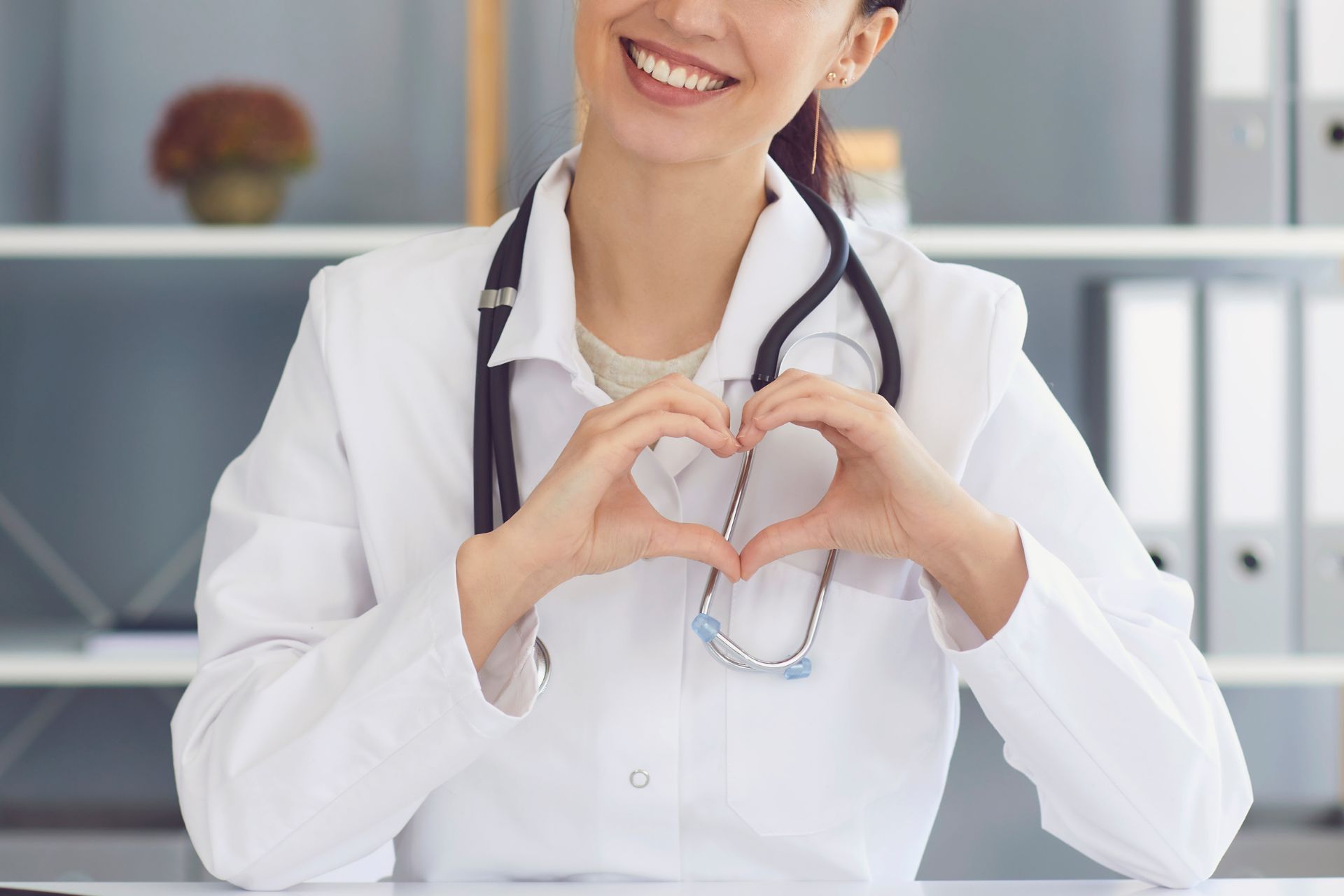 Doctor in white coat makes a heart shape with hands, smiling. Stethoscope around neck.