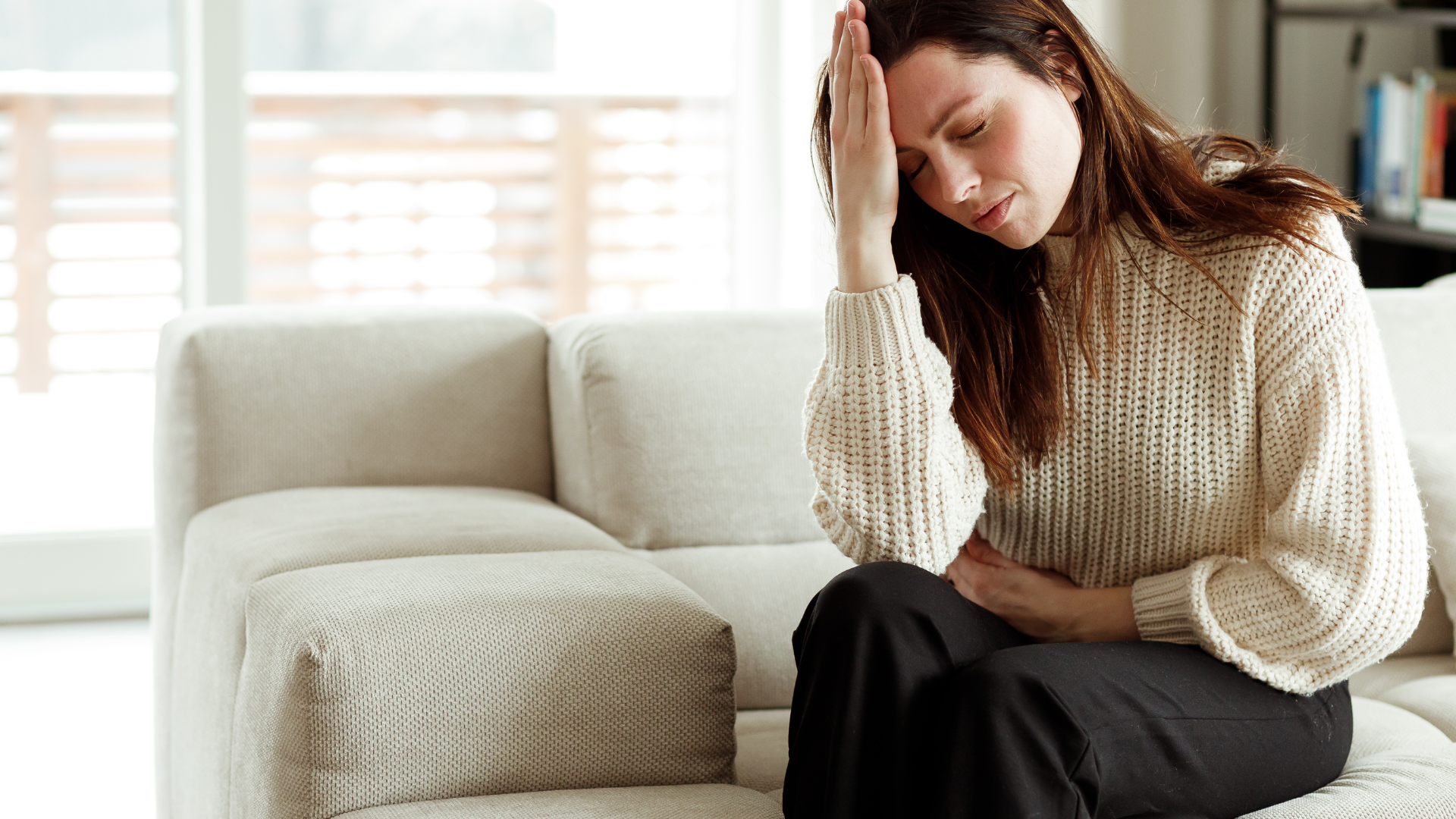 Woman sitting on a couch with hand on forehead, looking down. Interior setting, neutral colors.