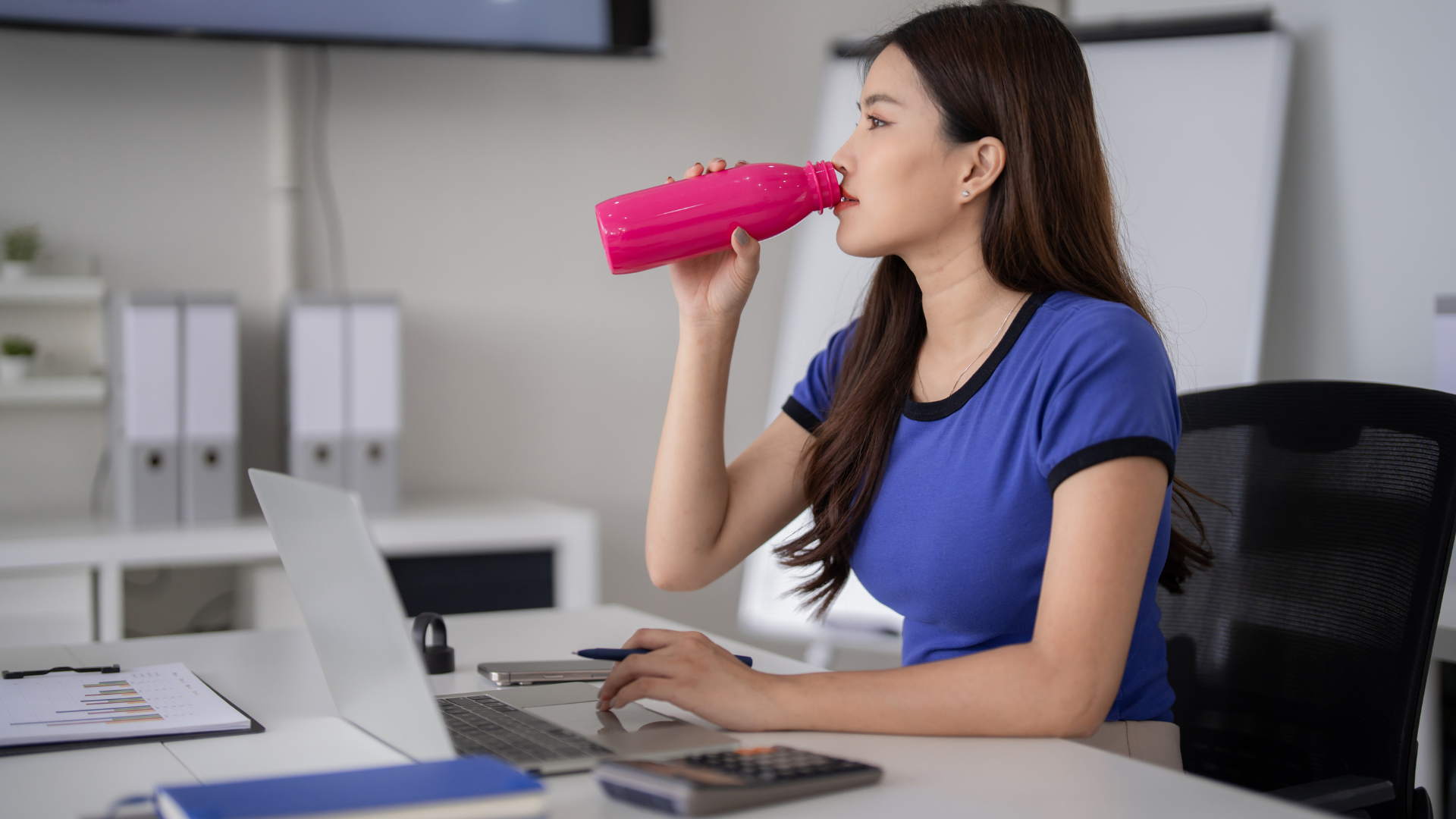 Woman drinking from a mug while leaning against a wooden table with a laptop, neutral background.