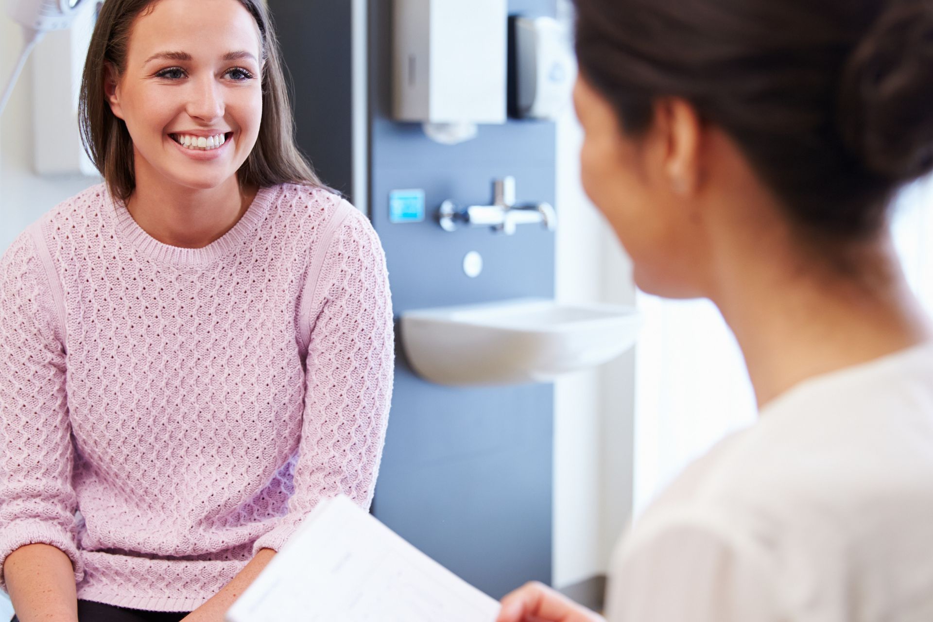 Woman in pink sweater smiling at person holding papers in a medical office with a sink and dispenser.