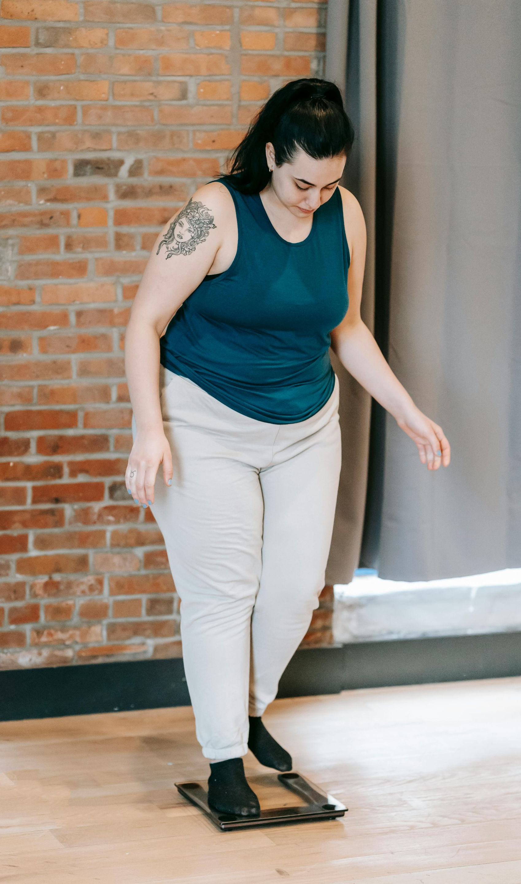 Woman in blue tank top and white pants standing on a scale in a room with a brick wall.