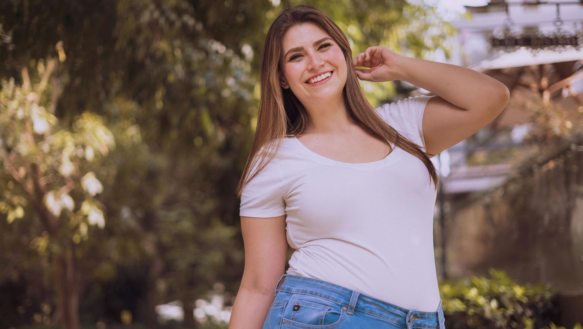 Smiling woman in white shirt and jeans, posing outdoors with trees in the background.
