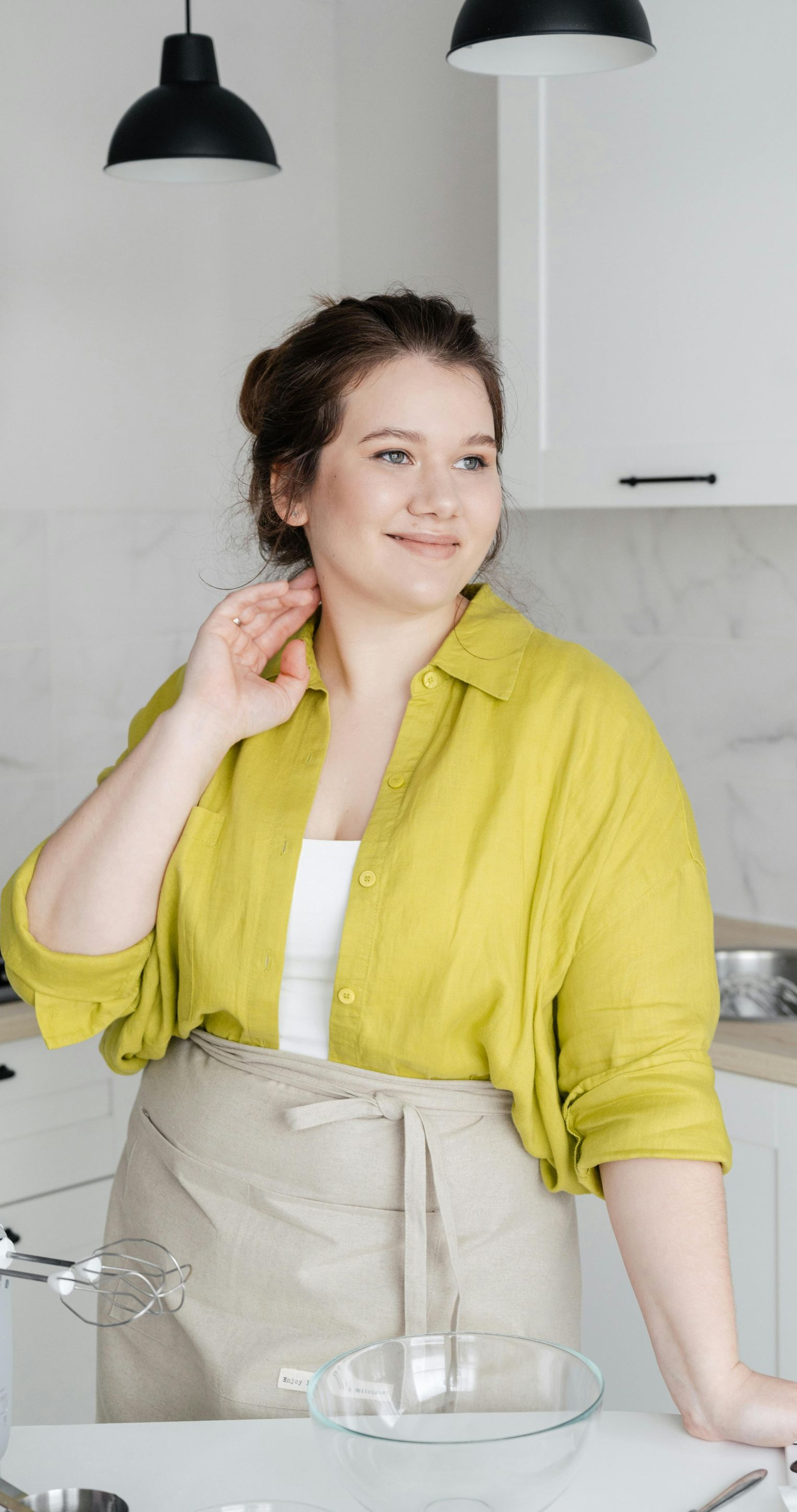 Woman in kitchen, wearing an apron, smiling, hand near neck. Bright yellow shirt, white top.
