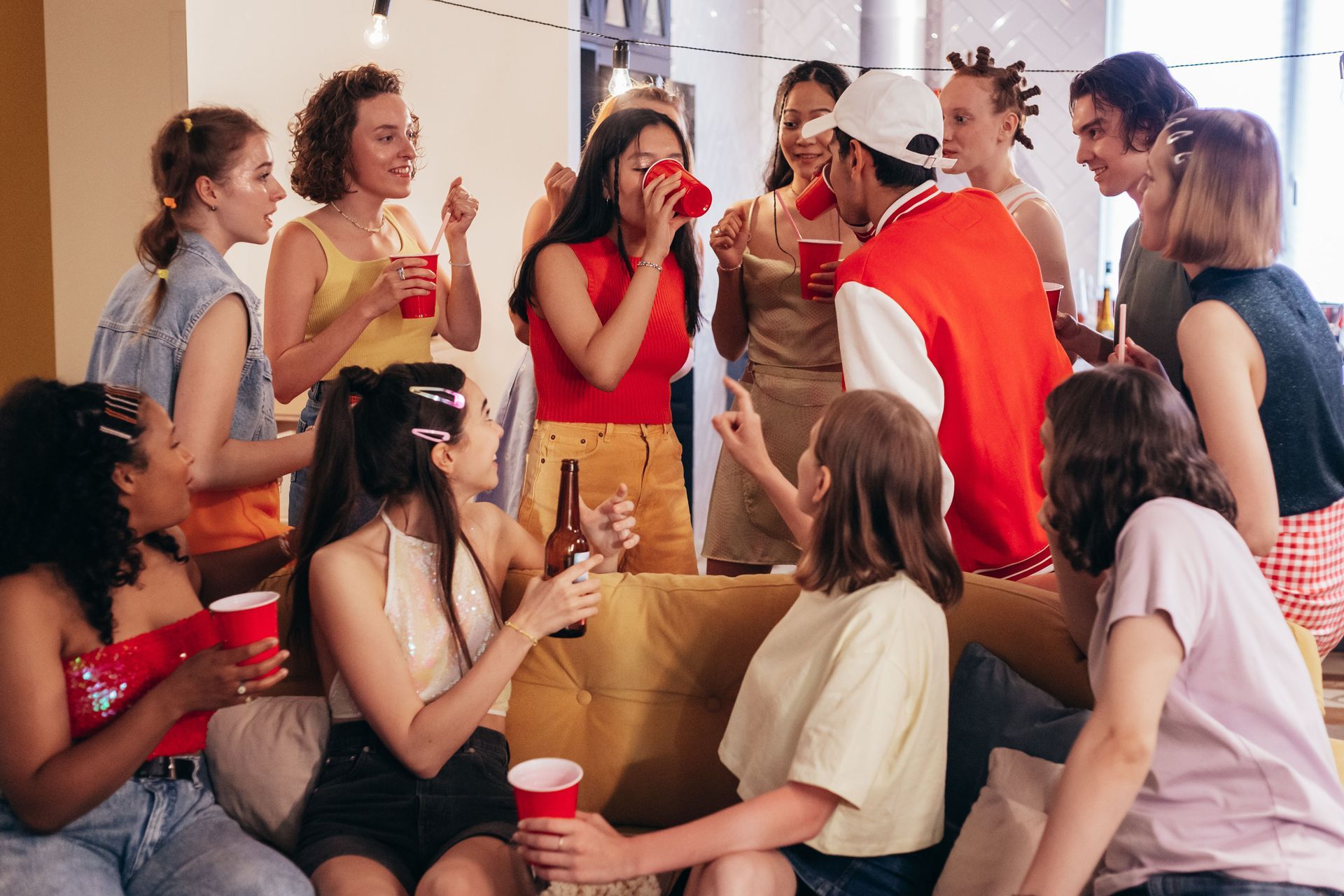 Group of people chatting and laughing at an indoor party, holding red cups and snacks around a couch.