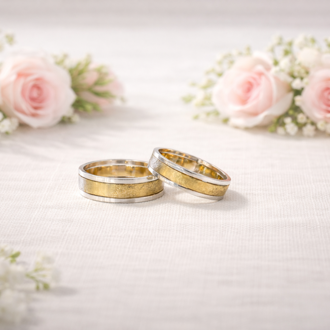 Twee bicolor gouden trouwringen op een wit kleed met zachtroze rozen op de achtergrond.