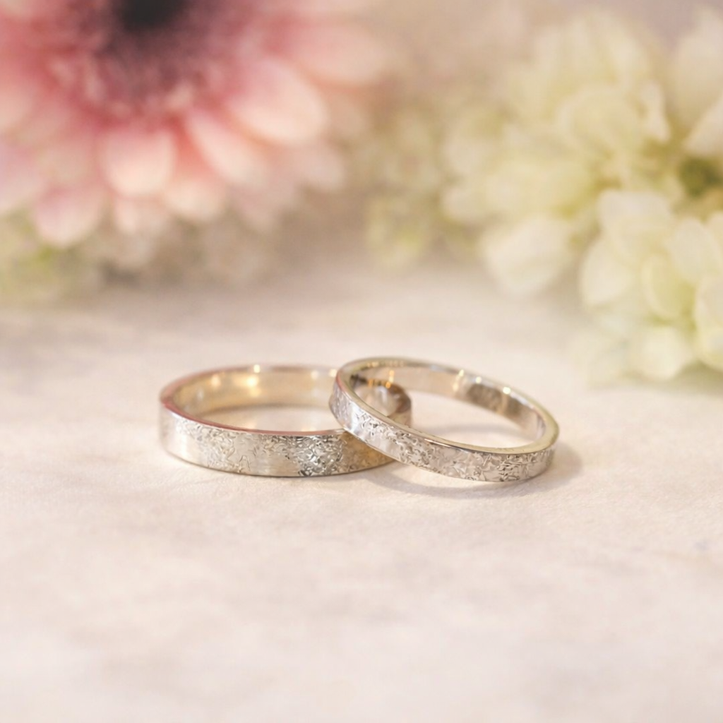 Twee witgouden ringen op een zachtwitte ondergrond met wazige roze en crèmekleurige bloemen op de achtergrond.