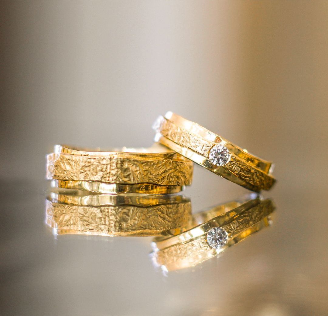 Twee gouden trouwringen op een reflecterend oppervlak, de ene tegen de andere aanleunend.itgouden