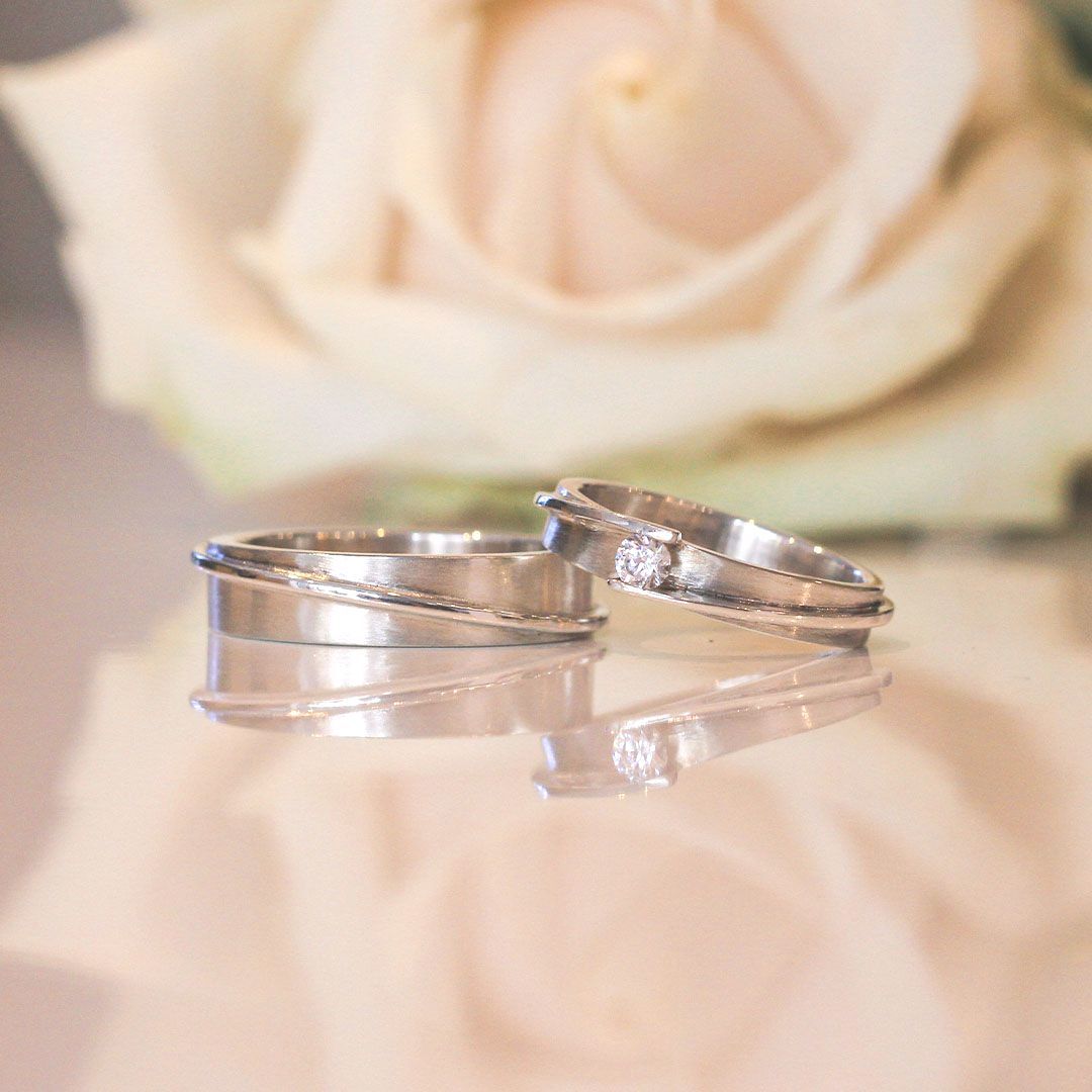 Twee witgouden ringen met diamant op een reflecterend oppervlak met een zacht gefocuste crèmekleurige roos op de achtergrond.