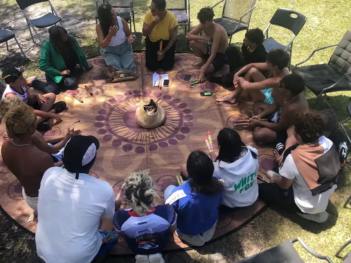 Group of people sitting in a circle on a rug outdoors, appearing to meditate or socialize.