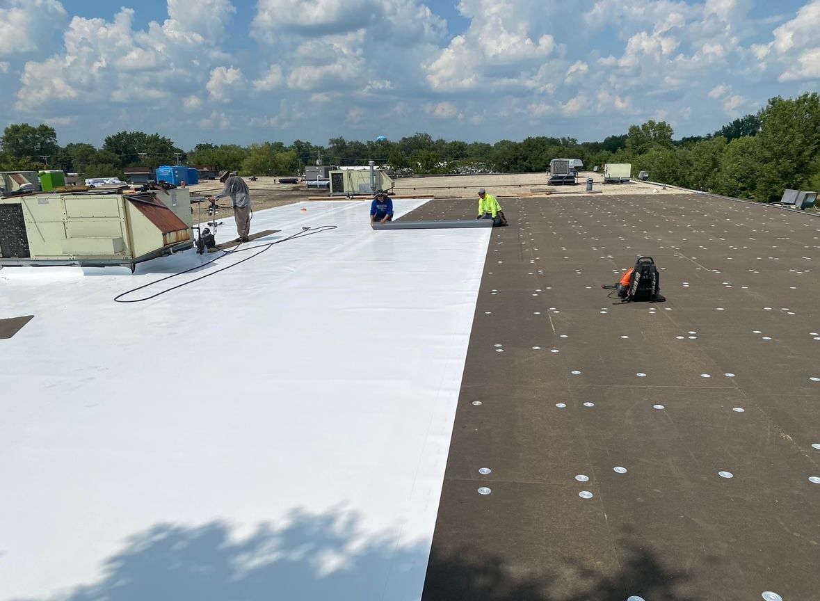 Workers installing a white roof membrane beside an unfinished brown roof under a cloudy sky.