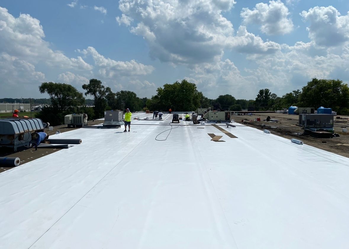 Flat rooftop covered with white membrane, workers and equipment under a cloudy sky.