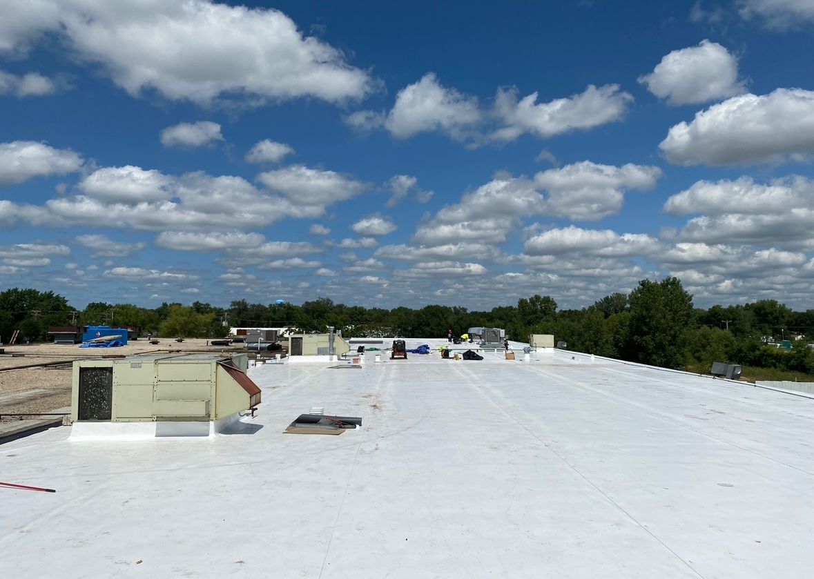 White rooftop under a bright blue sky with scattered clouds, vent structures, and distant trees and buildings