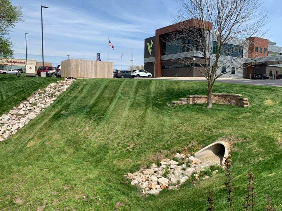 Green grassy slope with a culvert and rocky drainage area, near a building and fence.