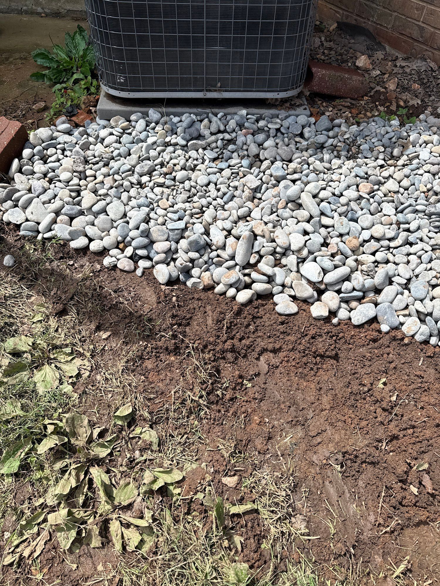 Gravel and dirt border an air conditioning unit. Dried weeds grow in the foreground.