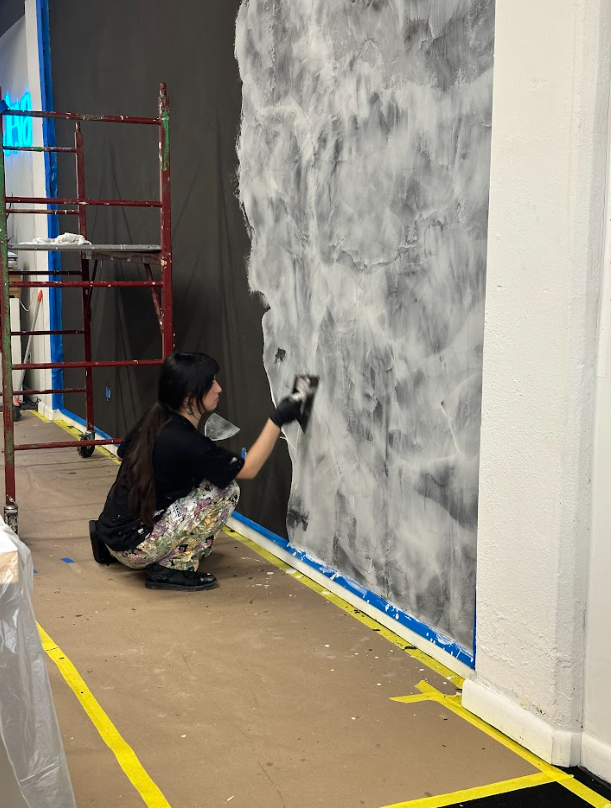 Person crouches painting a gray wall indoors near scaffolding and taped floor markings.