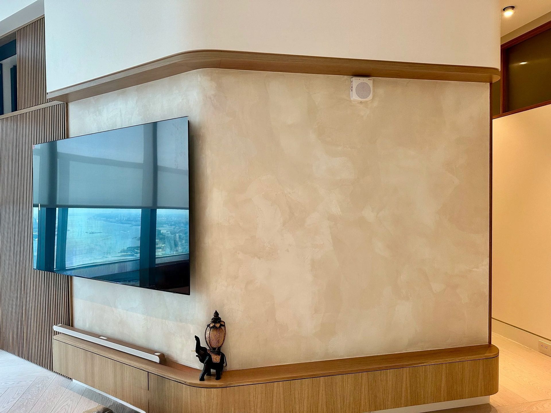 Corner wall with mounted TV and small shelf. Tan textured wall, wooden trim, small statue.