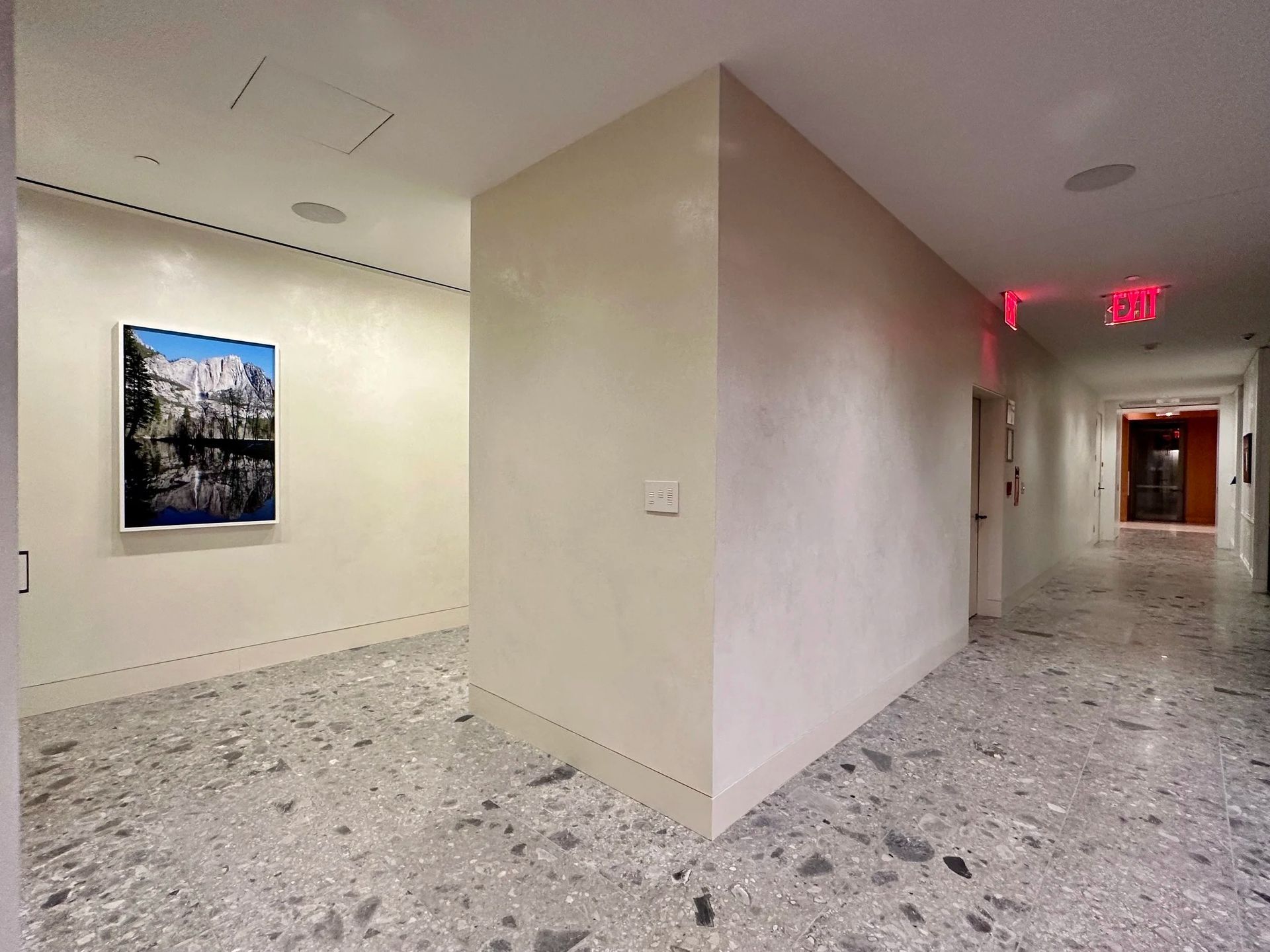 Hallway with terrazzo floor, artwork on the left, and exit signs at the end.